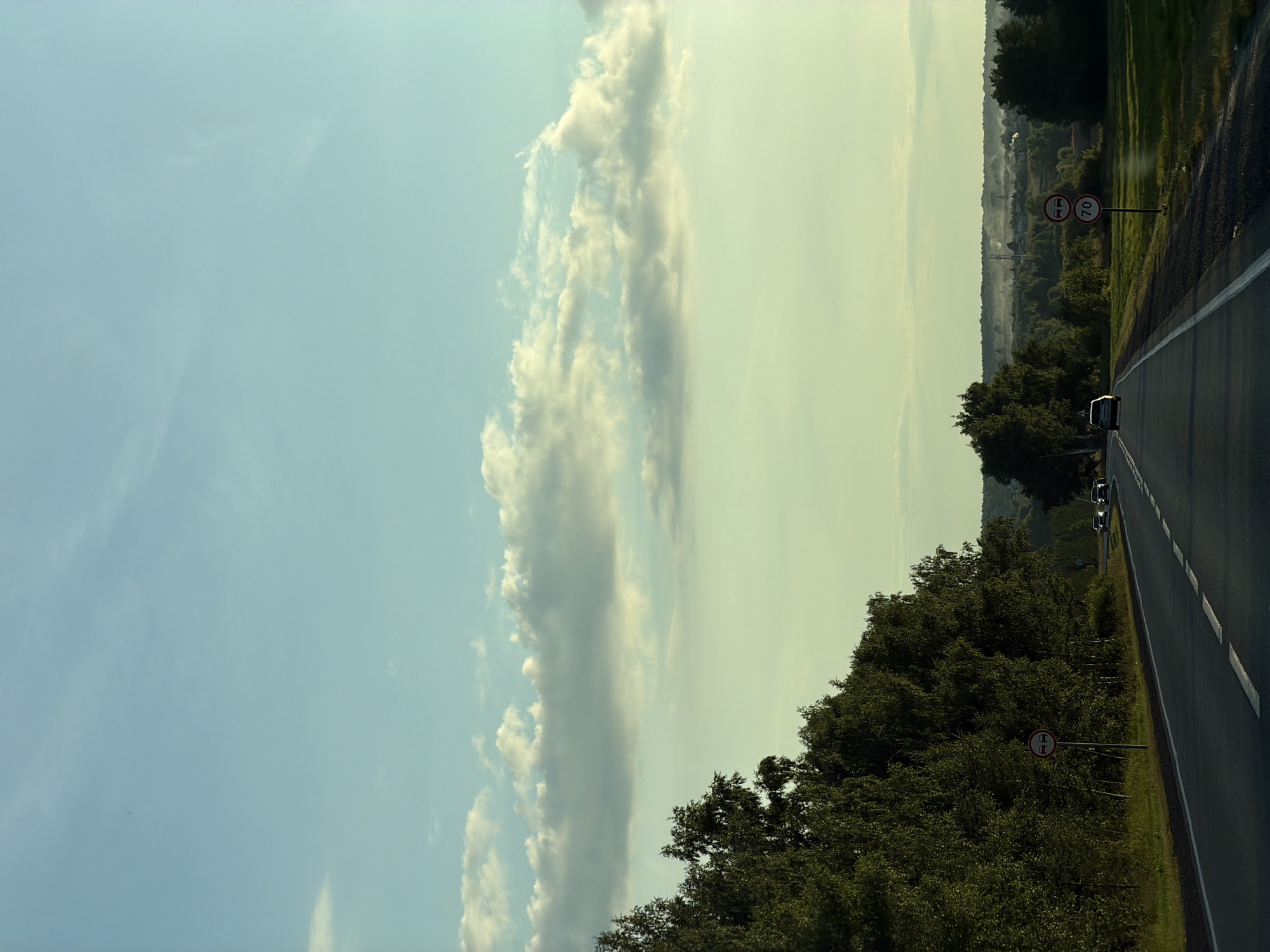 A car drives along a highway surrounded by trees. The sky is a pale blue with scattered clouds. In the distance, there is a village with smoke rising from chimneys. The image is captured from the perspective of someone inside the car. The scene suggests a rural setting.