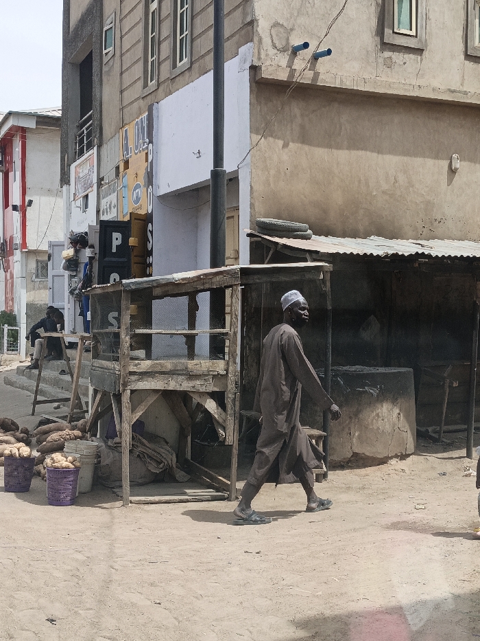 The image displays an outdoor street scene in Hotoro South, Nigeria, under bright daylight, with compacted dirt serving as the ground surface. In the foreground, a dark-skinned male subject, wearing a dark brown tunic, a white cap, and black sandals, is observed mid-stride, moving from right to left across the frame. To the left of the central path, two purple plastic baskets filled with white root vegetables are placed on the ground, accompanied by a pile of larger brown root vegetables. A two-tiered, wooden stall with mesh sides occupies the central midground. Behind this stall, to the right, stands a structure with a corrugated metal roof, topped by two stacked tires. Adjacent to this structure is a large, circular, concrete or earthen installation. In the background, a second male subject, dressed in dark clothing, is seated on wooden steps near a building on the left. The background consists of several multi-story buildings exhibiting varying architectural styles and finishes, along with a prominent black utility pole and overhead wires.
