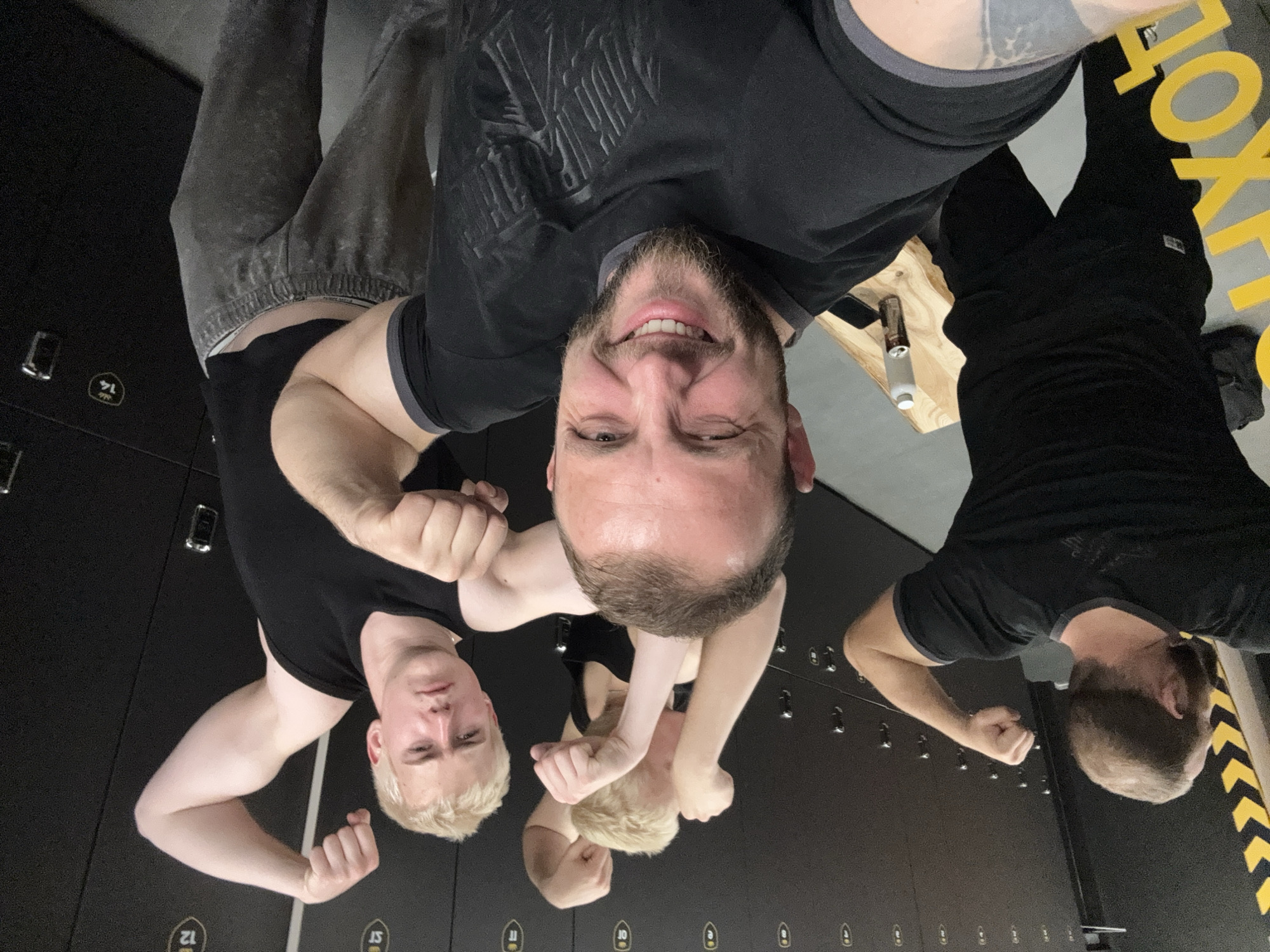 The image captures a group selfie of four men in what appears to be a locker room or gym changing area. The perspective is a wide-angle, low shot, making the central figure prominent and seemingly closer to the camera.
In the center, a man with a beard and dark hair smiles broadly and winks at the camera, holding it to take the selfie. He wears a black t-shirt with a subtle embossed design. To his immediate left (viewer's left), another man with blonde hair and a serious expression flexes his bicep while wearing a black tank top. Partially visible behind him is a third man, also with blonde hair, flexing his arms. To the right, a fourth man, seen from the back and side, also has a beard and is flexing his bicep, wearing a black t-shirt and showing a tattoo on his arm. The central man also has a visible tattoo on his arm. One of the men is wearing dark grey sweatpants.
The background features a row of dark grey or black lockers, some with numbers visible like "14", "11", "12", and "10". There are also small symbols next to some numbers, possibly "ST". To the right of the central man, a light-colored wooden surface serves as a bench or table, holding two bottles (one white, one metallic) and a mobile phone. Further to the right, a black wall or floor section features a yellow and black striped hazard pattern, and part of a large yellow word is visible, possibly "FOXT..." or "BOX...", suggesting a brand or gym name.
The overall mood is casual and playful, with the central figure's wink and smile, typical of a post-workout or social gathering in a fitness environment.