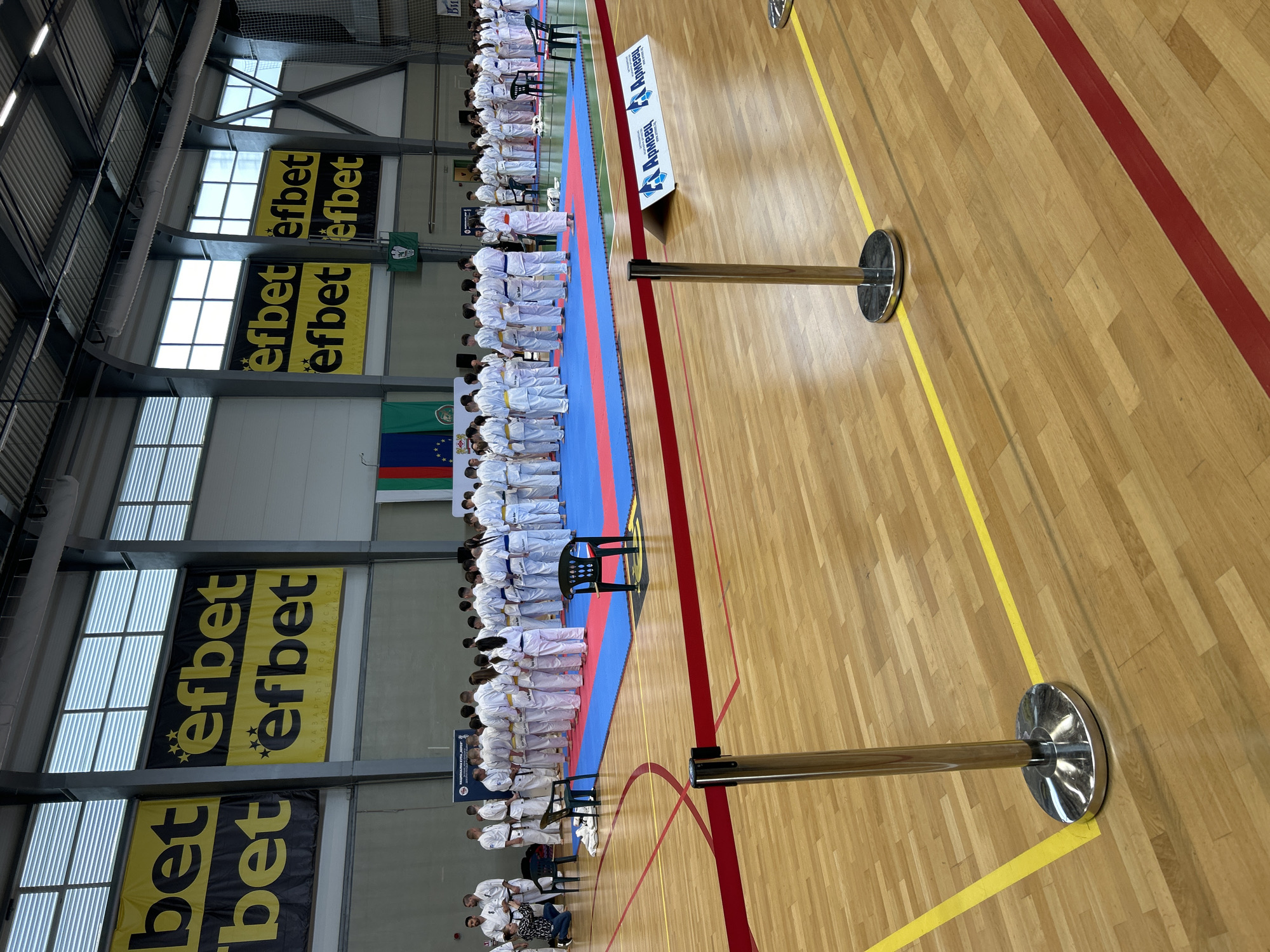 The image depicts a karate competition in a gymnasium in Stara Zagora, Bulgaria. A large group of karate students dressed in white uniforms and varying colored belts are standing in rows on a blue and red mat, seemingly in preparation for or following a match. The gymnasium has a wooden floor marked with red and yellow lines. Banners with the word "efbet" are visible above the students. The Bulgarian flag is also displayed. Chrome crowd control stanchions stand on the edge of the mat. A table, chairs, and additional people can be seen at the edge of the frame, potentially related to the event's organization or audience. The gymnasium has windows near the ceiling and a high ceiling with a structural framework.