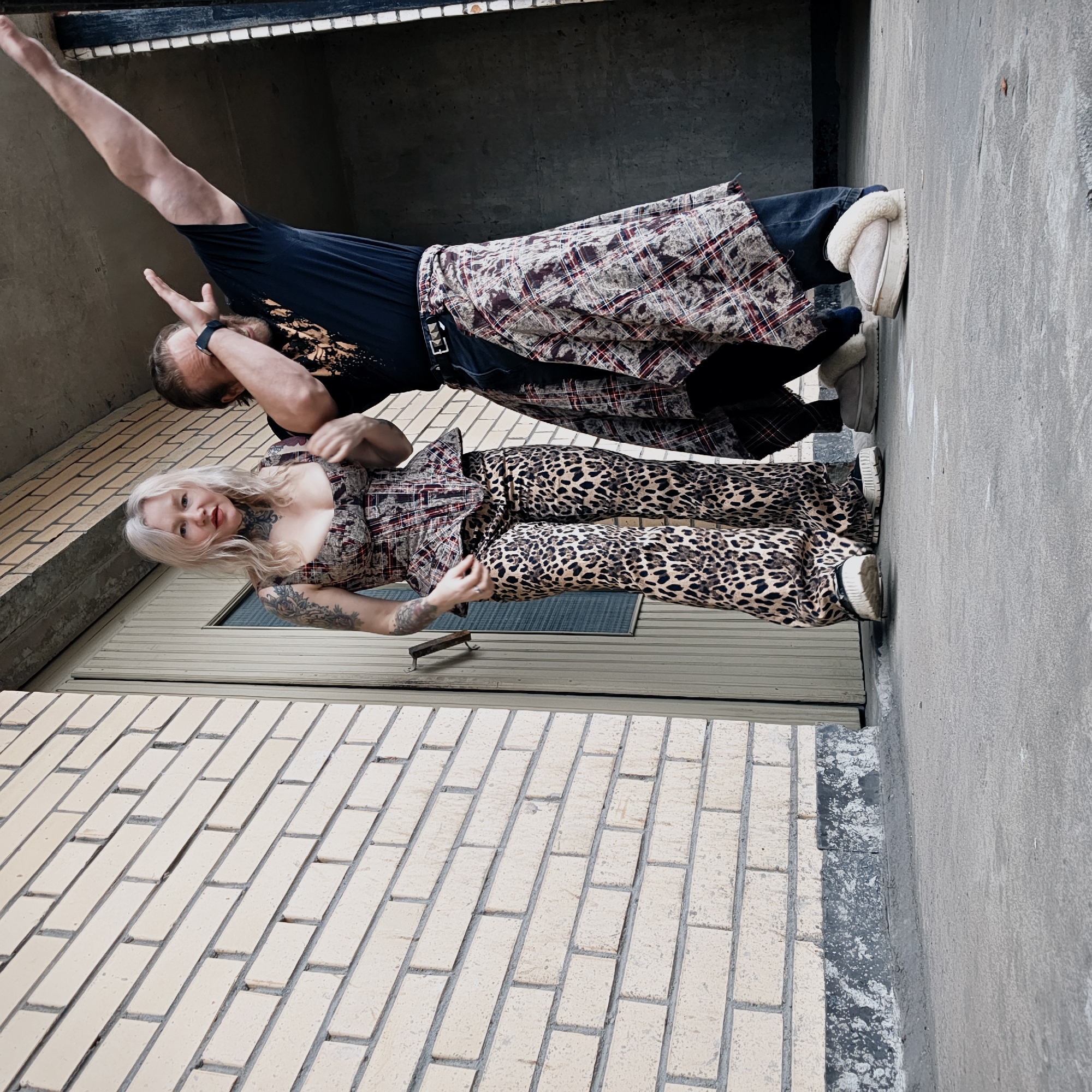 The image captures two individuals, a woman and a man, posing on a concrete surface against a light-colored brick wall and a dark concrete recess in Saint Petersburg, Russia. The photograph is taken from a low angle, looking upwards at the subjects.
The woman, on the left, has light blonde hair, tattooed arms and neck, and is wearing red lipstick. She is dressed in a plaid-patterned, corset-style top with a peplum, leopard print flared pants, and light-colored sneakers with thick soles. Her left hand is positioned near her chest, and she maintains eye contact with the camera.
The man, on the right, has darker hair and a beard, and wears a watch on his left wrist. He is attired in a black t-shirt with a light-colored graphic, dark trousers, and beige fur-lined slip-on shoes with thick soles. A plaid shirt is tied around his waist, secured by a black studded belt. He is captured mid-action, with his right arm extended upwards and bent at the elbow, and his left hand partially obscuring his face.
The immediate background to the left features a light beige brick wall with grey mortar, exhibiting some discoloration and a dark, patchy foundation section. A vertical, light-colored door frame or panel is partially visible behind the woman. To the right, the background transitions into a dark concrete alcove or underpass. The ground beneath the subjects is a textured, light grey concrete slab.