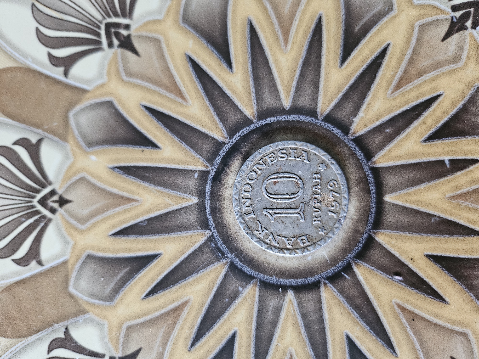 An Indonesian 10 Rupiah coin dated 1979 rests on a decorative tile. The tile features a radial design with alternating brown and beige triangular patterns. The coin is centered in the image. The surrounding tile appears to be part of a larger patterned surface. Jasinga, Indonesia is mentioned as a location, though the connection to the image is not explicit.
