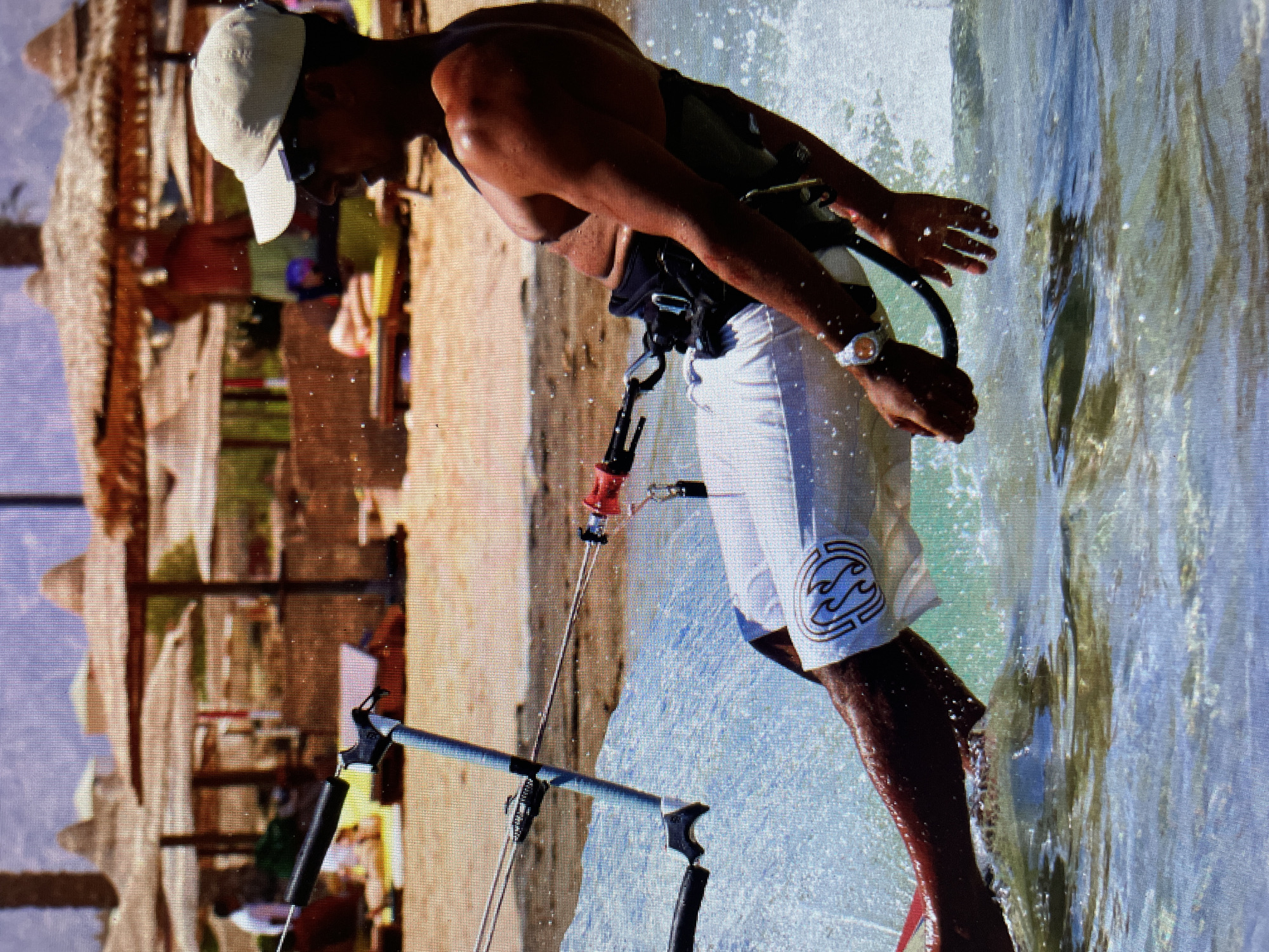 A man is kitesurfing in an area with shallow, clear water, likely a beach. The location could be Bacong, Philippines. The man is wearing a white cap, a black harness, and white shorts with a logo on the side. He is holding onto the kite control bar, and water is splashing around him. In the background, there is a sandy beach with beach umbrellas and possible other people.