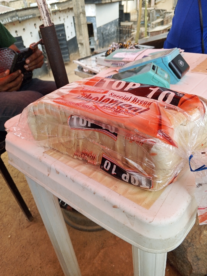 The scene is an outdoor commercial setting in Gwong, Nigeria. A plastic-wrapped loaf of "TOP 10 Special Bread Wholemeal" is centrally positioned on a white, scuffed plastic table. The bread wrapper displays "Special Bread" and "Wholemeal" text in red and black. To the left, a person with dark skin wears a green shirt and dark trousers, holding a black, textured object with both hands. A dark metal pole stands vertically in front of this individual. To the right of the bread, partially obscured by the bread itself, another person is visible from the shoulder up, wearing a blue garment. Behind the bread, on the same table, two light-blue electronic devices (possibly POS terminals or weigh scales) are present, along with papers and other small items. The ground beneath the table is reddish-brown earth. In the background, multiple low-rise buildings with light-colored walls and dark accents are visible, suggesting an urban or market environment.