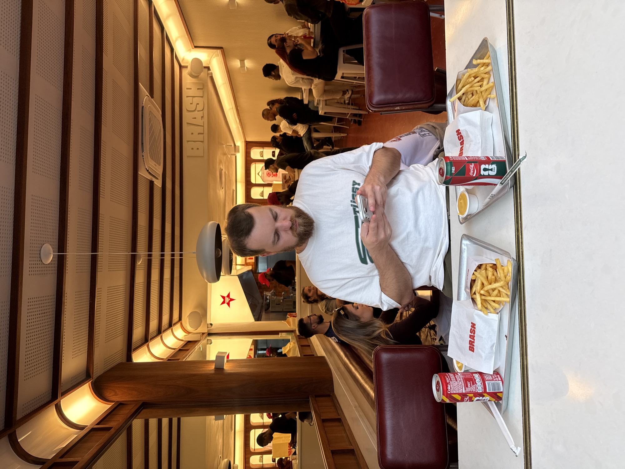 The image depicts the interior of a restaurant called "Brash" in Cape Town, South Africa. A man with a beard is in the foreground, using his phone. In front of him is a tray containing fries, a "Brash" branded bag, a small container of sauce, a Coca-Cola can and paper straw wrappers. A woman with long hair sits beside him. The restaurant has rows of seating with other customers visible in the background, mostly seated at tables. The interior design features brown wood elements, panelled walls, and modern lighting fixtures. The walls have "BRASH" spelled out in large letters. A red star design is visible on a white wall in the background.