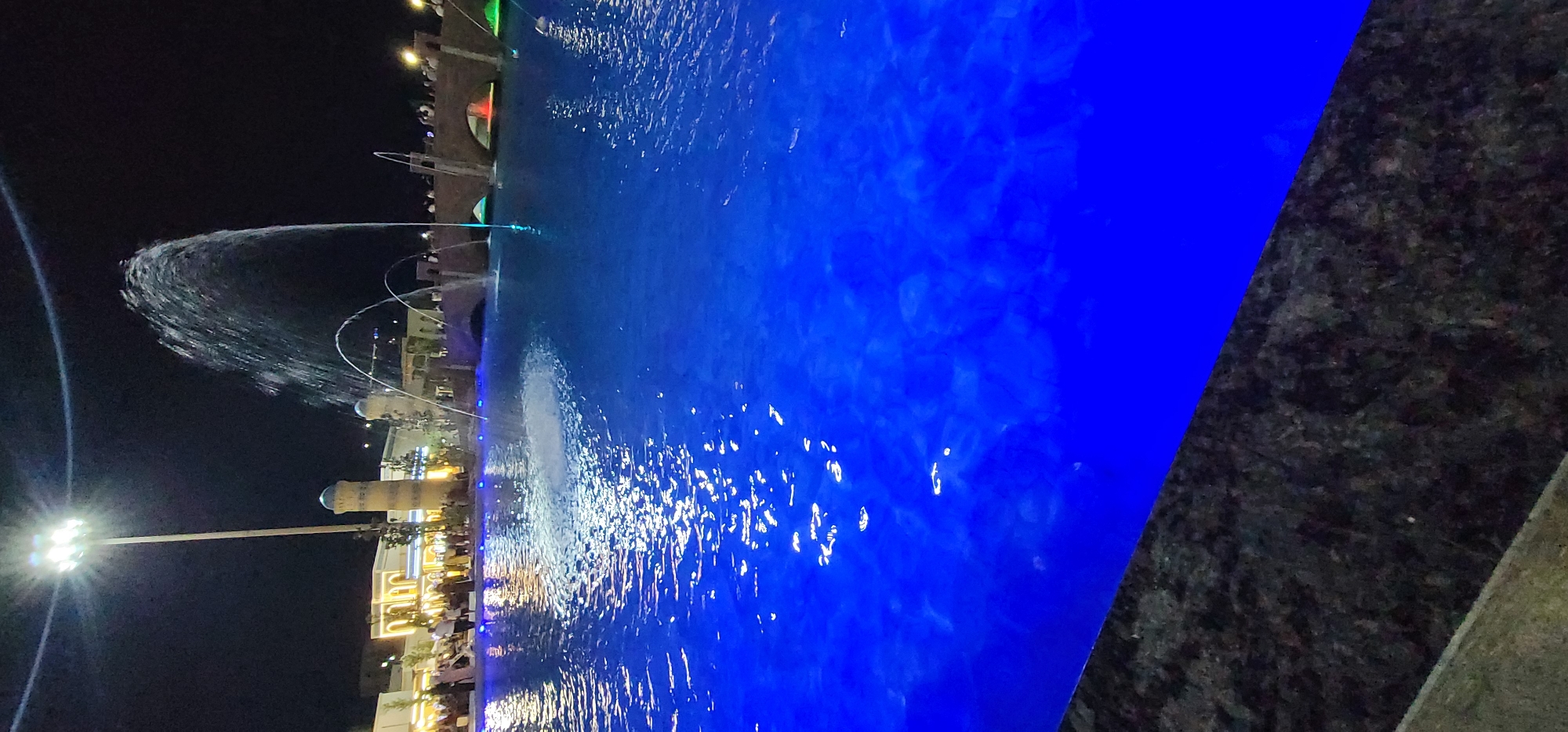 This is a nighttime outdoor photograph taken in the city of Shakhrisabz, Uzbekistan, specifically at the Nadir Divan-Beghi Madrasah complex. A large, illuminated pool of water dominates the foreground, glowing with an intense blue light from underwater fixtures. The water's surface reflects the light, creating shimmering patterns. In the background, a large fountain shoots water high into the air, illuminated by the surrounding lights.
Behind the fountain, a bridge with arched openings spans across a body of water, with people gathered along its edge. To the left of the fountain, the distinctive minarets and facade of the Nadir Divan-Beghi Madrasah are visible. The building is lit by warm, yellow lights, highlighting its architectural details. A tall, modern light pole with multiple floodlights stands prominently near the madrasah, casting bright white light. Several trees are scattered around the area, and many people are walking around, suggesting a public gathering or event. The overall atmosphere is lively and festive, with the combination of water features, illuminated architecture, and people creating a dynamic nighttime scene.