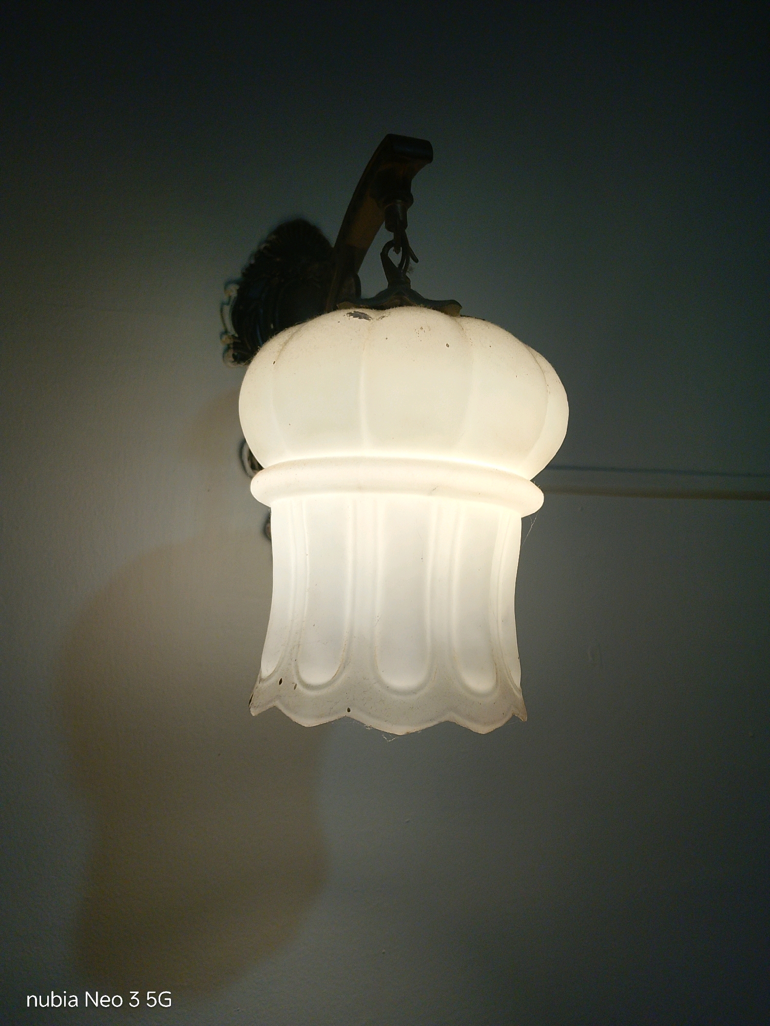 A close-up, low-angle shot captures an ornate, illuminated wall sconce hanging against a dark, textured wall. The sconce features a frosted glass shade with a ribbed, bell-shaped design and a fluted bottom, emitting a warm, diffused light. Above the shade, a dark metal fixture with decorative detailing is visible, attached to the wall by a chain. The background is a gradient of dark gray, with a subtle shadow cast on the left side of the wall by the sconce. The bottom left corner of the image displays the text "nubia Neo 3 5G". The image was taken in Tembilahan, Indonesia.