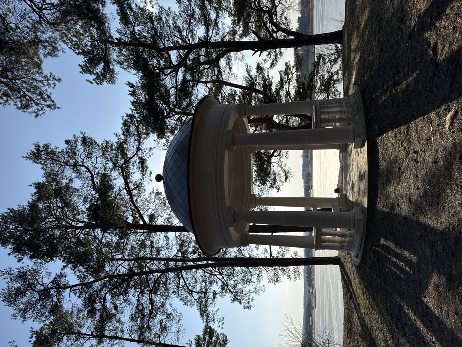 A white, circular, classical-style gazebo with a domed roof and balustrade stands on an elevated grassy area, overlooking a large body of water, in Падлабенскі сельскі Савет, Belarus. The gazebo is supported by multiple columns. Tall, slender pine trees frame the structure and the distant horizon, appearing largely silhouetted against a clear, light blue sky. The sun is positioned low behind the gazebo, creating strong backlighting and casting long, distinct shadows from the trees and gazebo across the dry, sparse foreground ground. The water's surface reflects sunlight. Distant structures are visible on the far shoreline. No individuals are present in the scene.