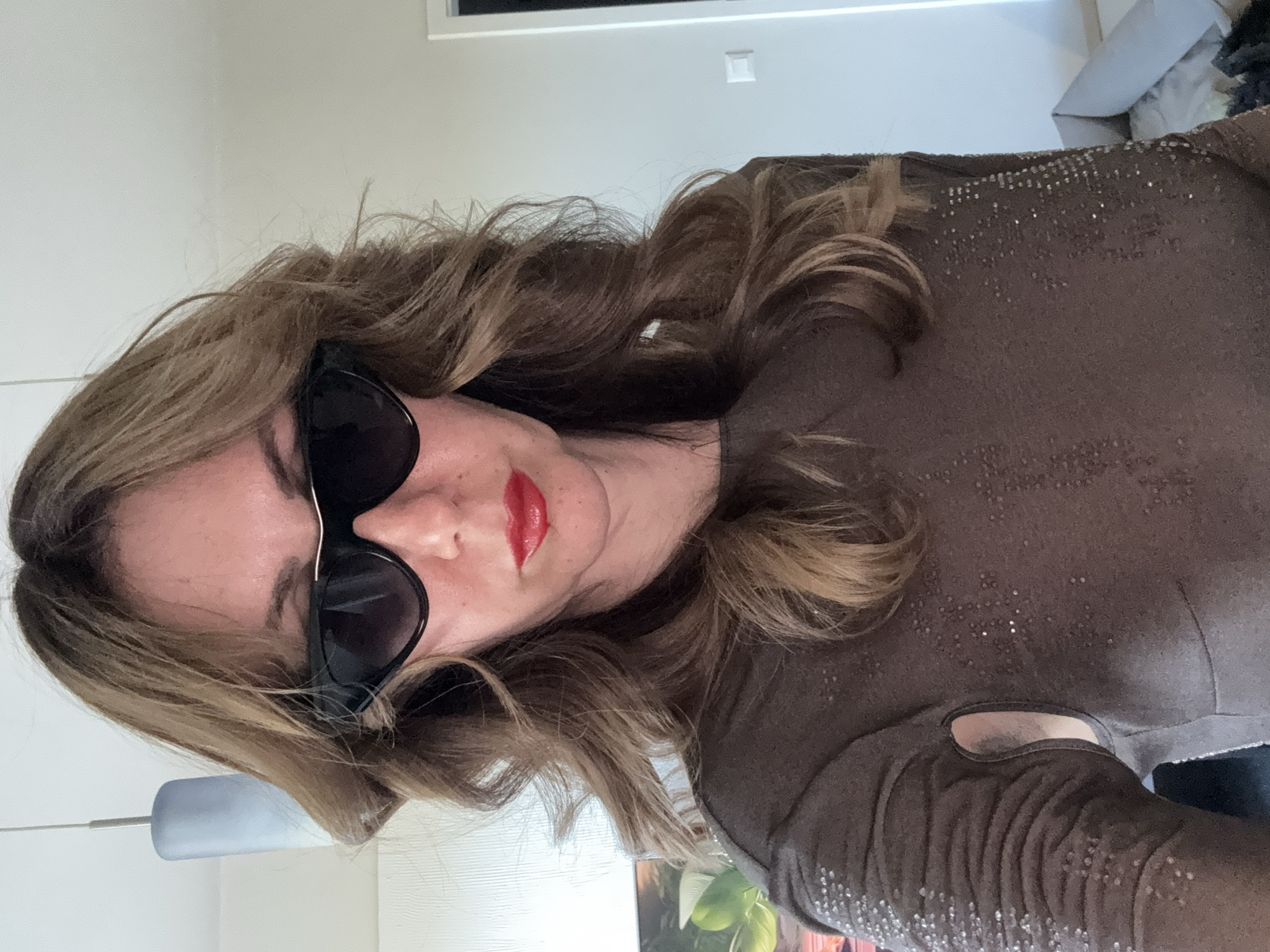 A woman with long, wavy brown hair and red lipstick is wearing sunglasses and a brown, sequined long-sleeved shirt with a cut-out on the shoulder. The background includes a white wall with a door and a light fixture. The location cannot be determined to be Sofia, Bulgaria from this image.