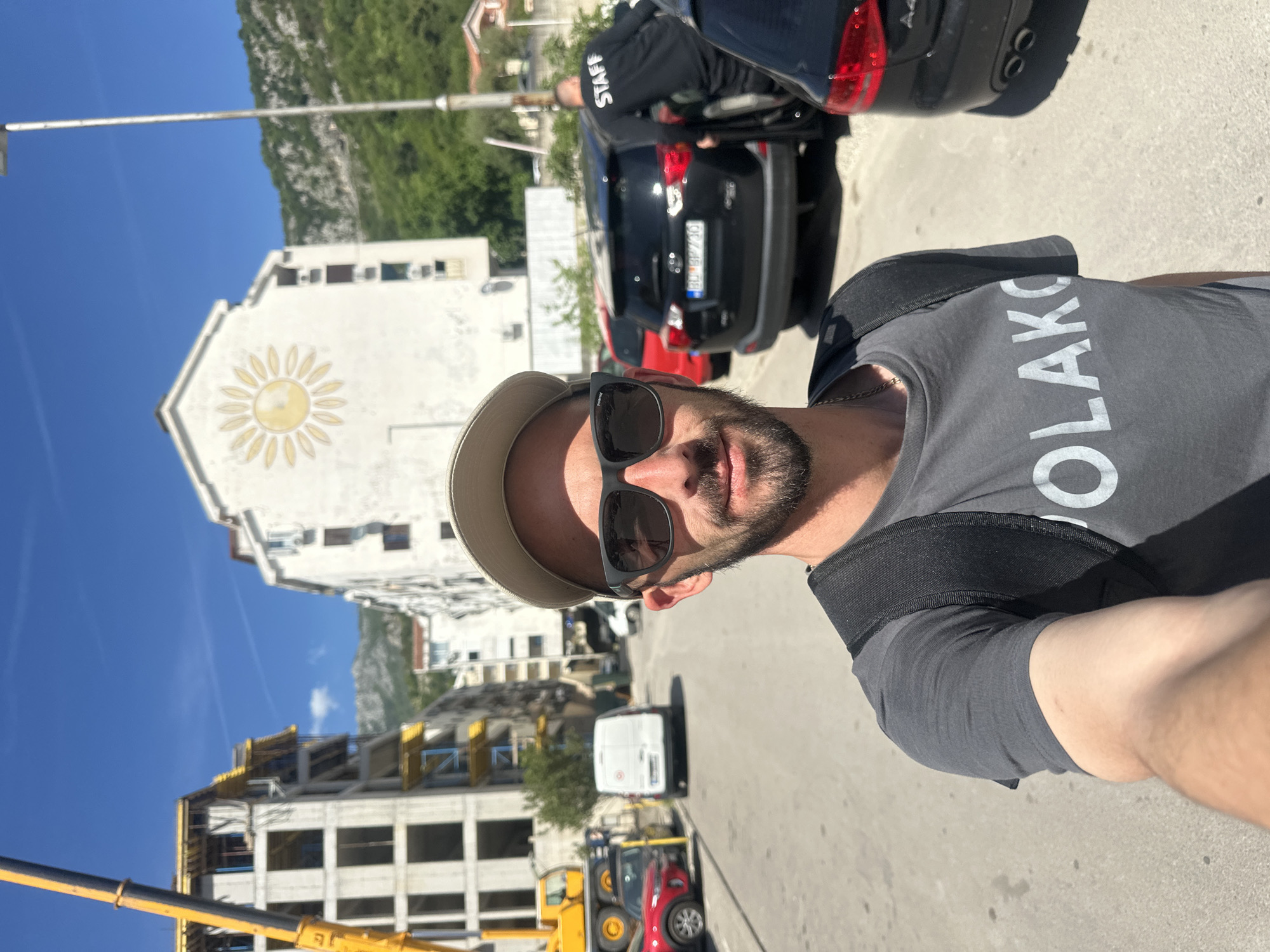 A selfie-style photograph captures a man in the foreground, turned slightly towards the camera, against a backdrop of a sunny town scene. The man has a beard, wears sunglasses, a light-colored baseball cap, and a dark grey t-shirt with a black strap or sling over his left shoulder. Visible text on his shirt reads "OLAKO".
Behind him, the scene unfolds under a clear blue sky. A prominent white building features a large, yellow, sun-like emblem painted on its facade. To the left, another building appears to be under construction, with yellow scaffolding or formwork visible. A yellow crane boom extends into the lower left of the frame.
Several vehicles are parked on a paved street, including a black SUV with a license plate "BU BP730", a red car, and a white van further down the road. Near the black SUV, a person wearing a black top with the word "STAFF" clearly visible on the back is seen interacting with the vehicle. In the far background, a large green mountain range rises under the bright sky. The clear light indicates it is daytime, likely late morning or afternoon, with sunny weather. The license plate's "BU" prefix strongly suggests the location is Budva, Montenegro, aligning with the provided context.