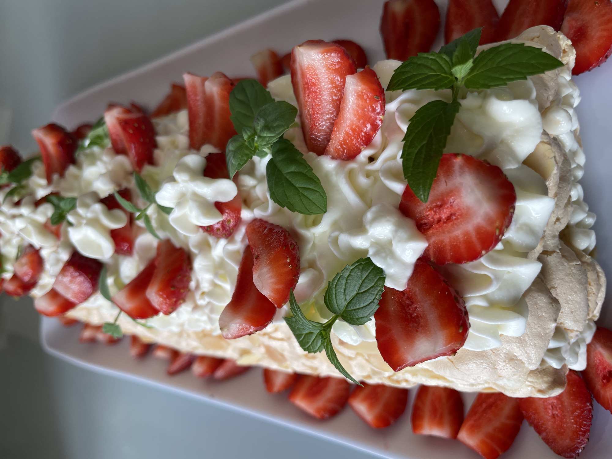 A close-up, overhead shot shows a long, rectangular pavlova cake topped with whipped cream, sliced strawberries, and fresh mint leaves. The cake is served on a white platter. The background is blurred and appears to be a light-colored, possibly indoor surface. There is no identifiable location within Saint Petersburg, Russia.