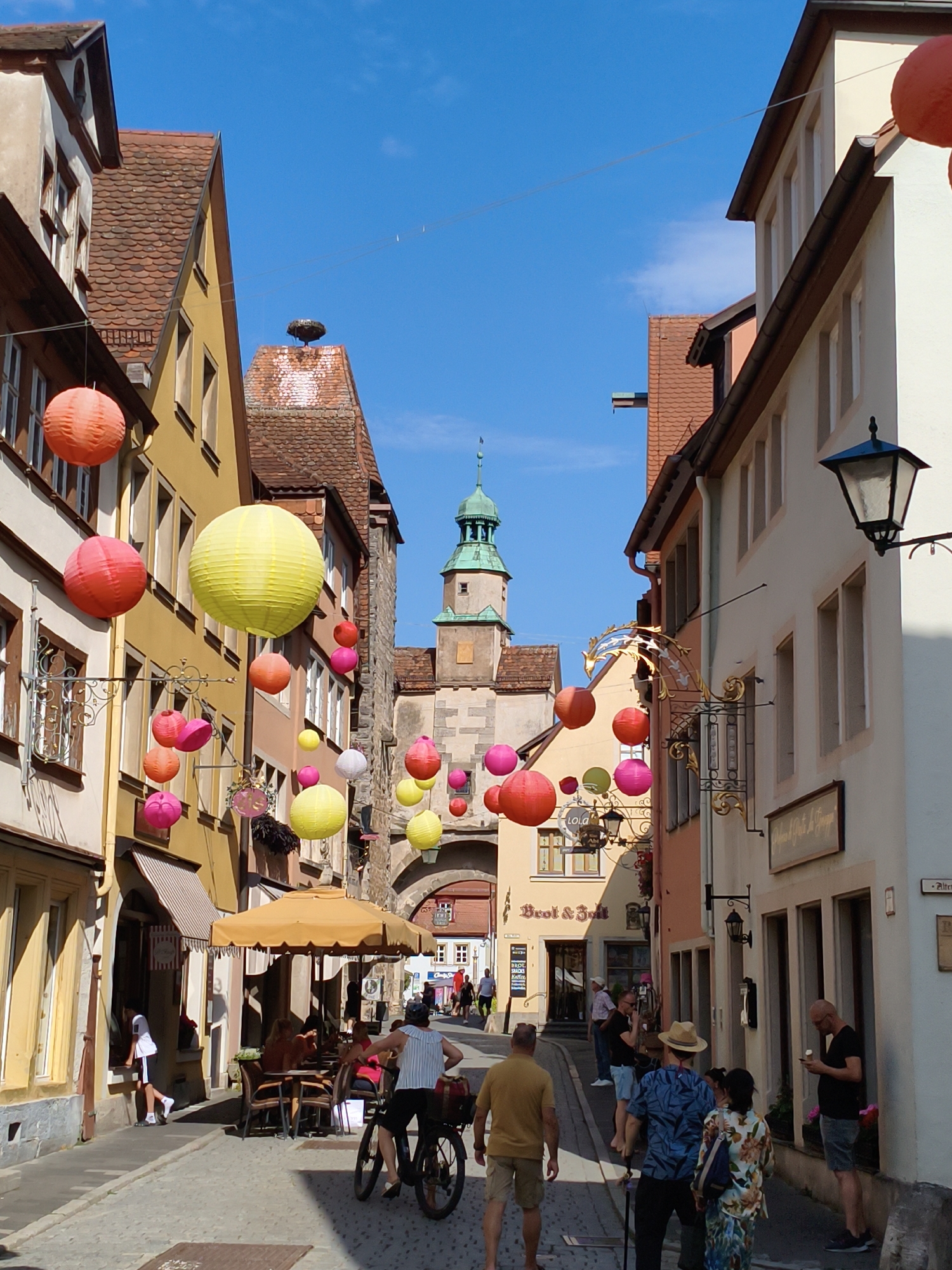 In Rothenburg ob der Tauber, Germany, a narrow cobblestone street is adorned with colorful hanging lanterns. The street is lined with buildings featuring traditional German architecture, including red-tiled roofs and cream-colored facades. In the foreground, a person on a bicycle is viewed from behind, wearing a striped shirt and dark shorts, with a basket attached to the rear of the bike. To their left, a man in a yellow t-shirt and khaki shorts walks away from the camera. Further down the street, people are seated at tables outside a cafe under a large yellow umbrella. In the background, a medieval tower with a green spire stands against a clear blue sky, and a stork's nest is visible atop a nearby building. The overall atmosphere suggests a vibrant and picturesque tourist destination.