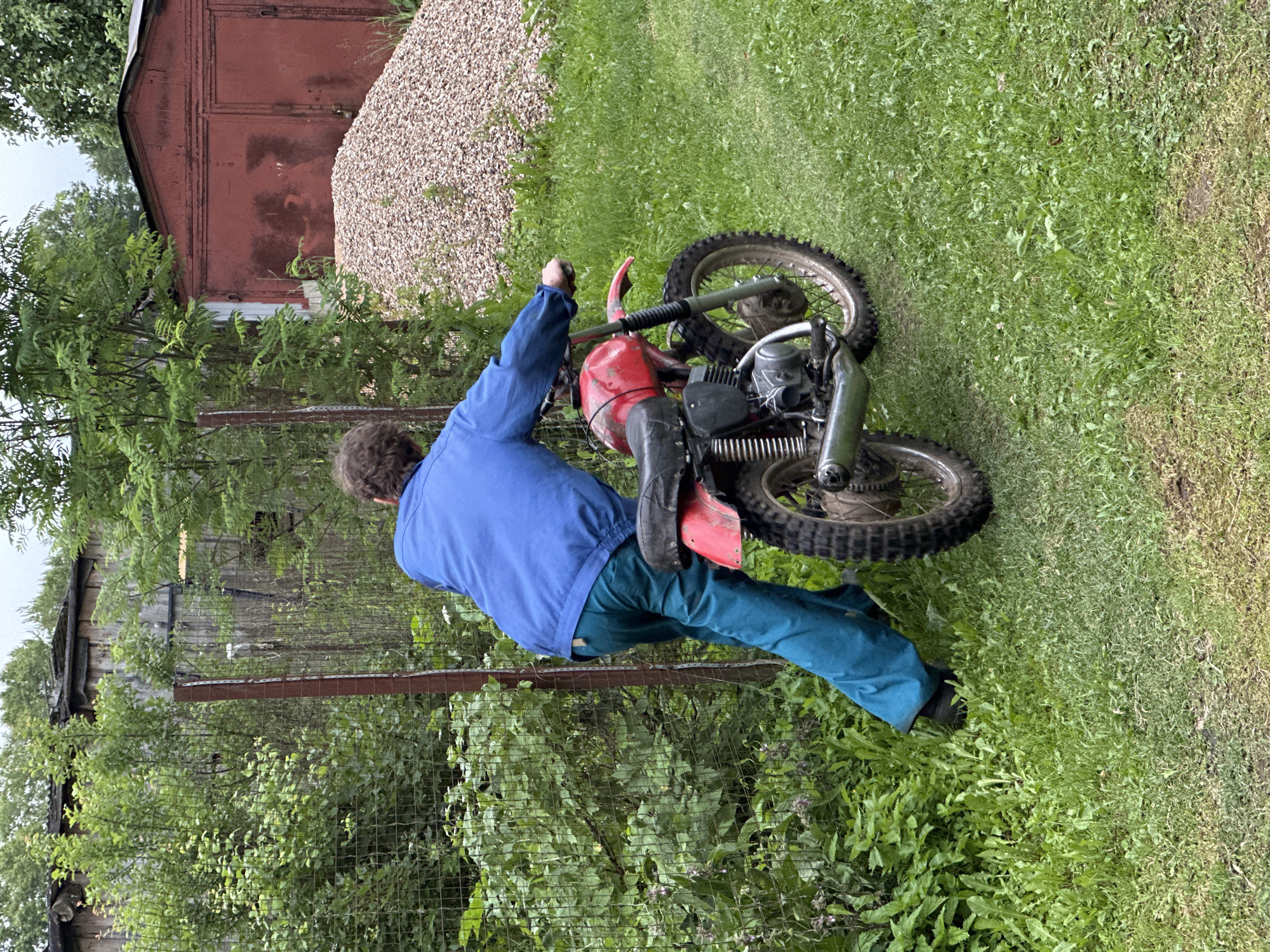 A person in a blue jacket and teal pants is pushing a red dirt bike on a grassy area. The person is facing away from the camera, with their right hand on the handlebars. The dirt bike has a black seat and silver exhaust pipe. In the background, there is a chain-link fence, a wooden fence, a pile of gravel, and a red garage. The scene appears to be in a rural or semi-rural setting, possibly in Piriutino, Russia, given the presence of a typical Russian garage and the overall atmosphere. The lighting suggests an overcast day.