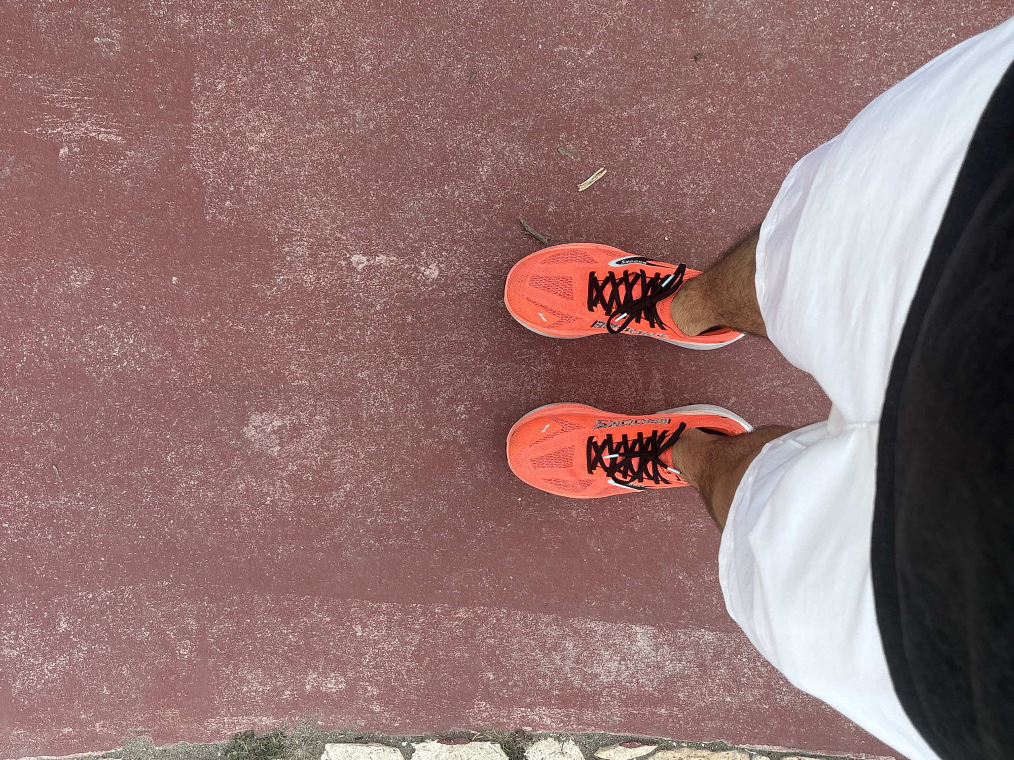 This top-down, high-angle shot captures a person's lower legs and feet on a reddish-brown textured surface, likely an outdoor running track or paved path. The person is wearing bright orange Brooks running shoes with black laces and white trim. The brand name "BROOKS" is visible on the side of the shoes. The person's legs are hairy, and they are wearing white shorts or athletic wear, with a glimpse of a black garment on the right side of the frame. The reddish-brown ground is speckled with lighter spots, suggesting wear or a particular surface type. A few small pieces of natural debris, such as twigs, are scattered on the ground. At the very bottom edge of the image, parts of white stones or pavement and some green vegetation are visible, indicating the edge of the main surface. The lighting suggests it is daytime. The scene implies the person is engaged in an activity like running, jogging, or walking.