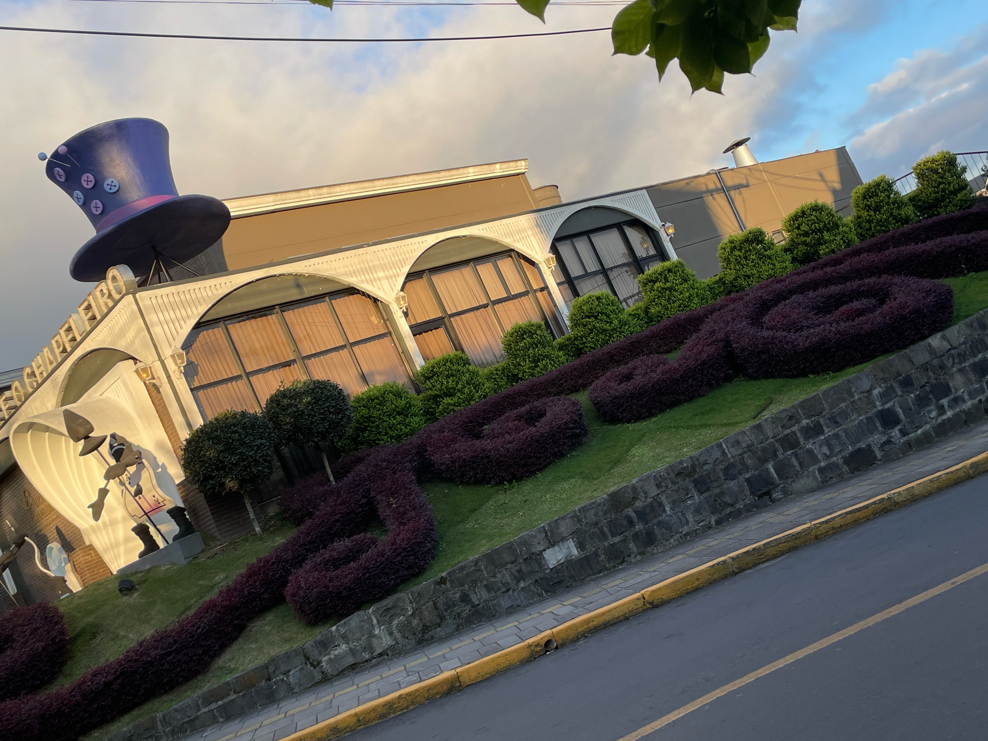 The image shows the exterior of a building in Gramado, Brazil, with a large purple top hat adorned with buttons and pins perched atop the entrance. The building features arched windows with golden curtains, and a sign with white lettering that reads "CASA CHAPELFEIRO". In front of the building, dark red bushes are sculpted into ornamental shapes on a grassy incline. A stone wall separates the landscaped area from a road with yellow curbs and a gray asphalt surface. The sky is partly cloudy, with hints of blue and the warm glow of sunset. No people are visible in the scene.