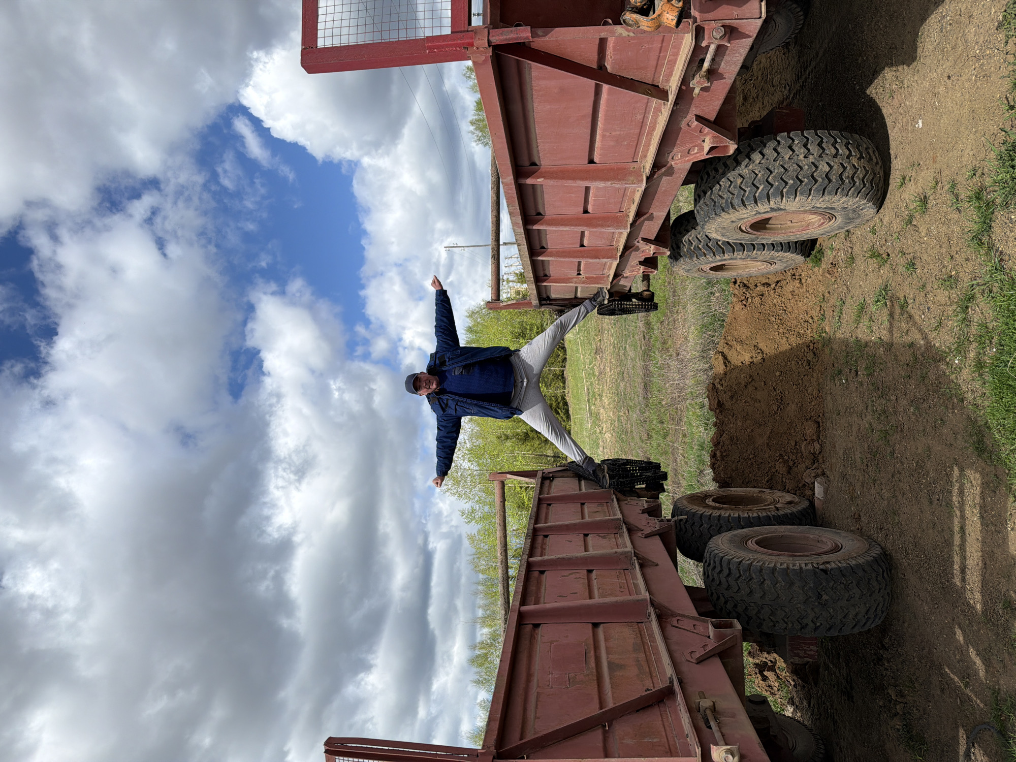 A man stands with a wide stance on the edges of two red trailers, arms outstretched, under a sky with scattered clouds. The location appears to be an outdoor area near Фоки, Russia, with greenery and bare earth visible.