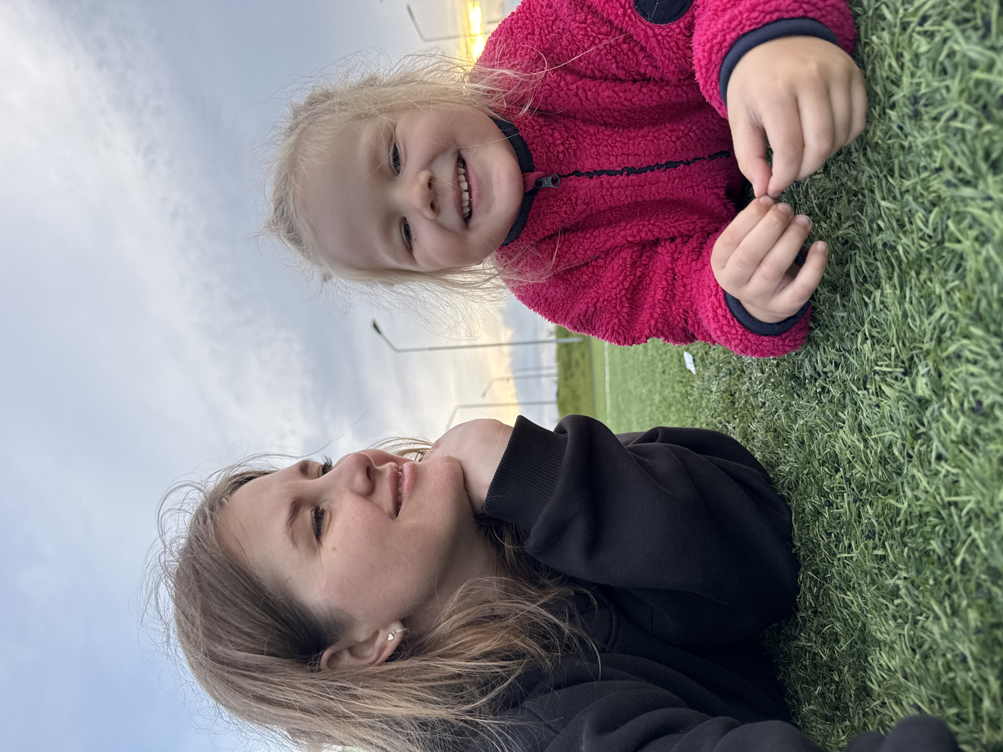 An adult female and a young child are lying on artificial green turf in an outdoor setting in Ферма, Russia. The female, positioned on the left, wears a black hooded sweatshirt and lies on her stomach, resting her chin on her left hand. She has light brown hair, a heart-shaped earring in her left ear, and her gaze is directed towards the right with a faint smile. The child, positioned on the right, wears a bright pink fuzzy jacket with a dark zipper and collar, and lies on their stomach facing the camera. The child has blonde hair, is smiling broadly with visible teeth, and is holding a small, indistinct object between their fingers. In the background, under a partly cloudy sky transitioning to a yellowish hue on the right horizon, several tall streetlights are visible.