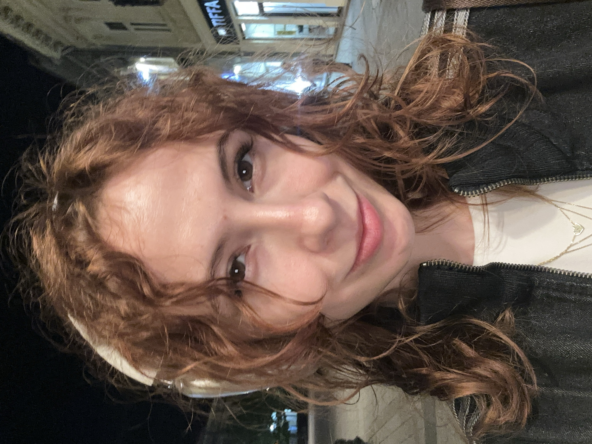 A close-up, rotated selfie shows a young woman with reddish-brown, wavy hair smiling warmly at the camera. Her head is tilted, and her dark brown eyes and a wide, closed-mouth smile are prominent. She appears to be wearing a dark, possibly denim, jacket with a visible zipper detail, over a light-colored top. A delicate gold necklace with a small heart-shaped pendant is visible around her neck.
The background is softly blurred, indicating an urban setting at night. Artificial lights from buildings or streetlamps are visible. A notable detail in the upper left background (when the image is viewed upright) is a partially visible illuminated sign that appears to read "TIFFA" or "TIFFANY," suggesting a store or brand name. The overall mood is casual and friendly, capturing a pleasant moment in what appears to be a city street in Novi Sad, Serbia.