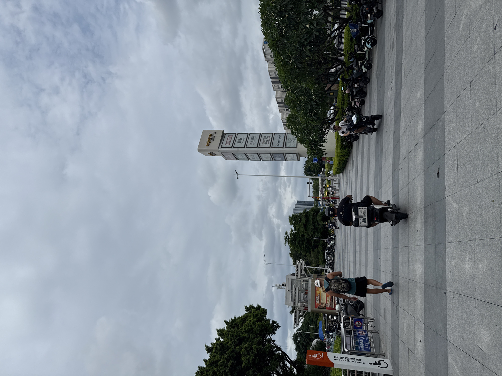 In Haizhu District, China, on a cloudy day, a paved pedestrian walkway is lined with parked scooters and motorcycles on the right side. A tall, multi-paneled signpost displaying various brands and text is prominently visible on the right, with lush green trees and bushes bordering the walkway. In the background, modern buildings with a consistent architectural style are visible. On the walkway, two individuals are riding scooters, and one person is walking away from the camera. A disabled access symbol is affixed to a signpost on the left side of the image.
