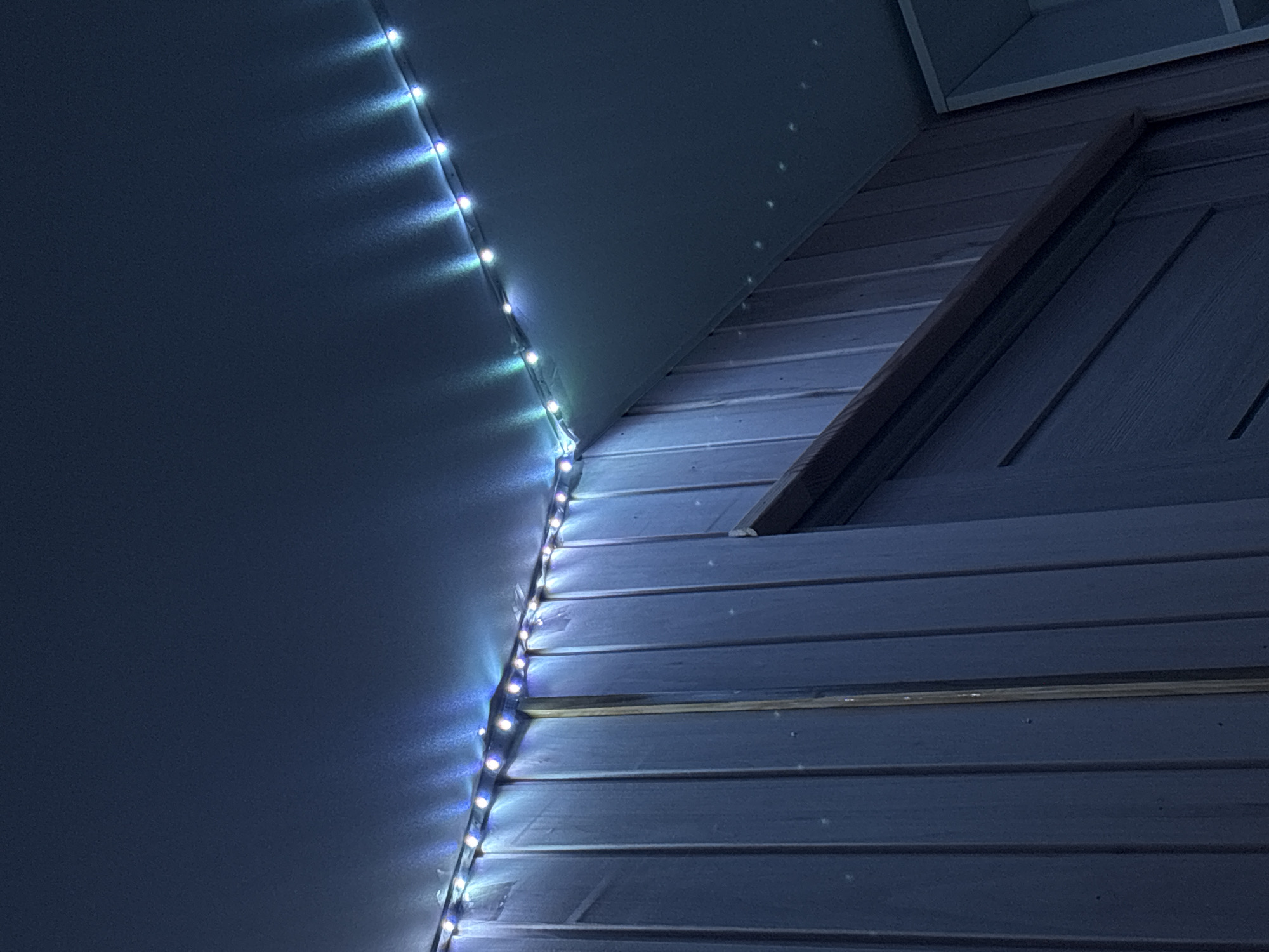 The image shows the exterior of a building at night. A string of blue and white LED lights is attached along the corner where the wall meets the roof. The building appears to be sided with horizontal planks. There are no people visible in the scene. The location is not verifiable as Rusalevka, Russia based on the image alone.