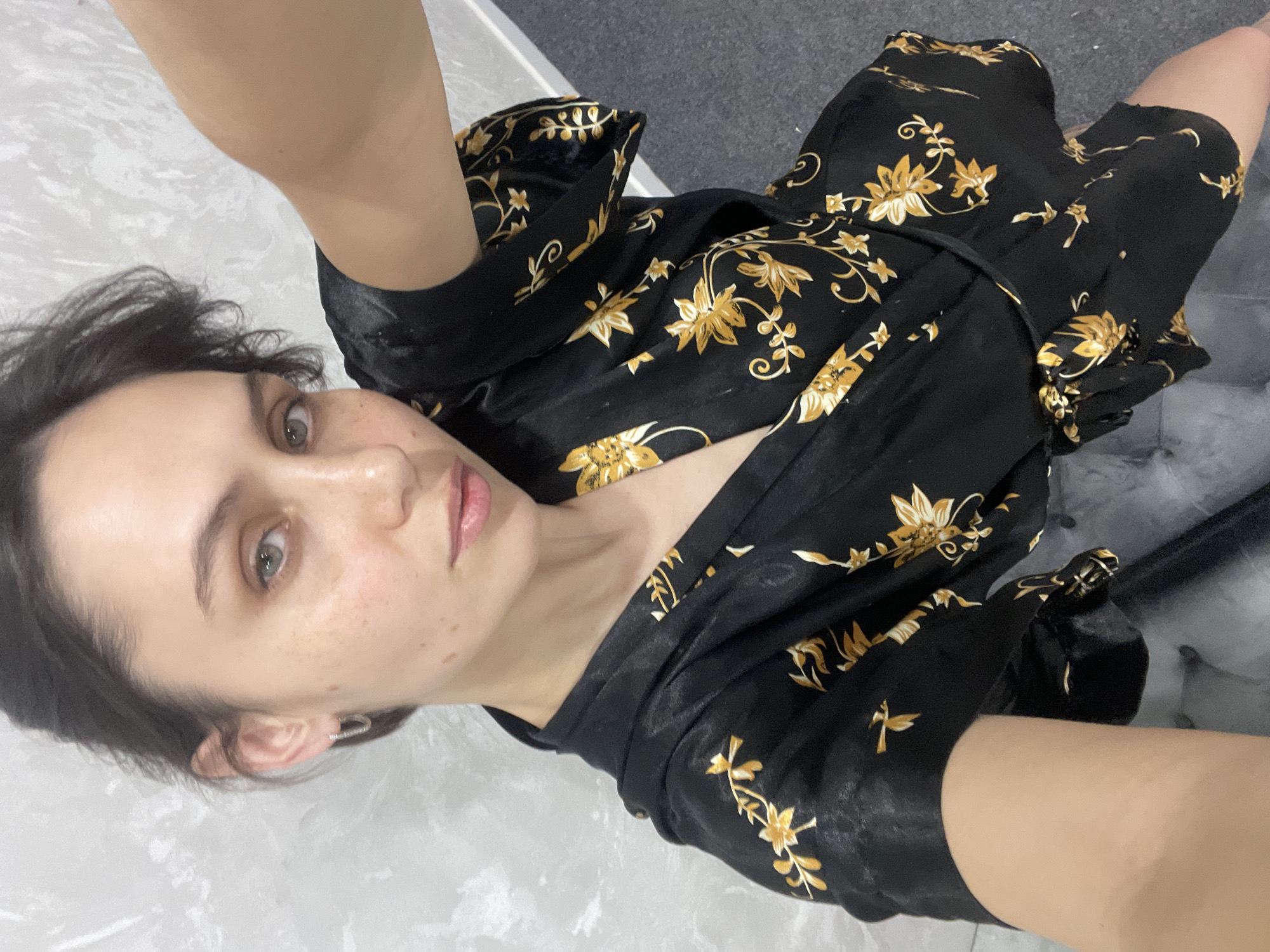 A woman with dark hair and fair skin is shown in a selfie. She has light-colored eyes and freckles on her face. She is wearing a black robe with a gold floral pattern. The background includes a white, marble-like surface and a gray, textured area. The image provides no information that can be used to identify this location as Izhevsk, Russia.