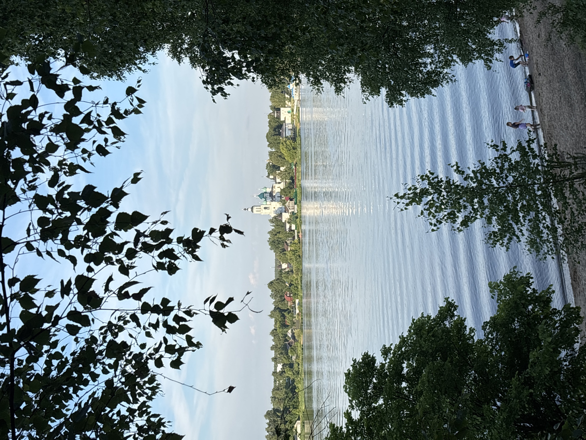 This is a photograph of a wide river with gentle waves, viewed from a tree-lined shore. In the distance, across the river, is a town with a prominent white and green church complex with multiple domes. The town is nestled amongst lush green trees, and houses are visible along the riverbank. The sky is a clear blue with some faint clouds. In the foreground, on the sandy shore, a few people are visible. Two children are playing near the water's edge, and another child is bending down to pick something up. There is also a dark-colored bag or mat on the sand. The scene is framed by the dark green leaves and branches of trees in the foreground. This scene is likely depicting the Volga River in the city of Yaroslavl, Russia, as it is a prominent feature of the city and the location of several notable church complexes.