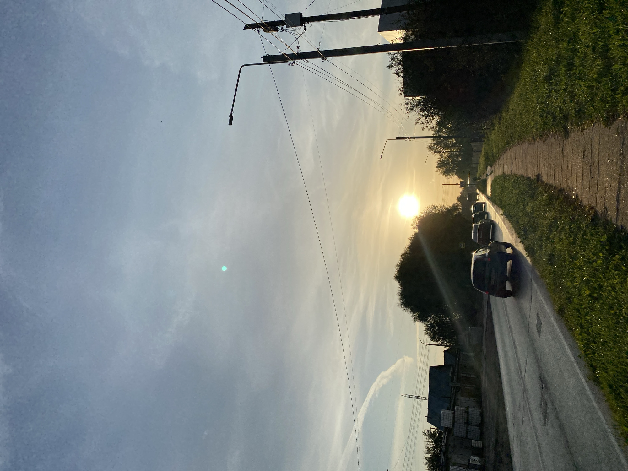 The image depicts a street scene in the city of Chaikovsky, Russia, during a sunset. A paved road runs diagonally from the lower left to the upper right of the frame. Several cars are traveling on the road, moving away from the viewer. To the right of the road is a concrete sidewalk, bordered by a strip of green grass. Beyond the sidewalk, a dense line of trees and bushes creates a dark silhouette against the sky. On the left side of the road, there are low-rise buildings, some appearing to be under construction or storage, with stacks of materials visible. Power lines are strung between utility poles that are visible on both sides of the street, with one prominent pole featuring streetlights. The sky is a pale blue with streaks of clouds, and the setting sun, a bright orb, casts a warm glow on the scene, with lens flare visible in the upper portion of the image. A contrail from an aircraft is visible in the upper left quadrant of the sky. The overall impression is of a quiet, early evening on the outskirts of a city.