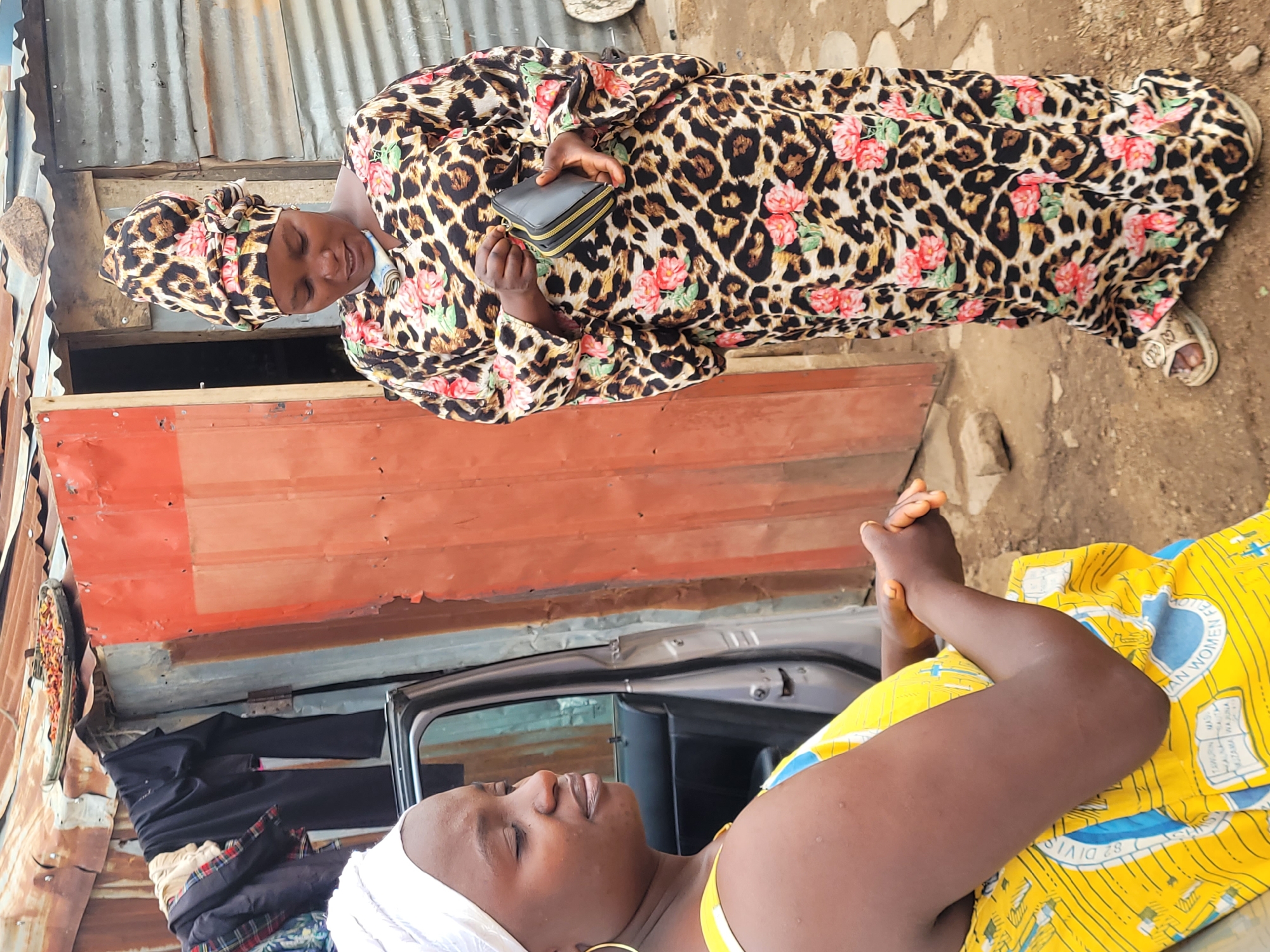 Two women are in an outdoor setting near buildings constructed of corrugated metal. One woman is standing, wearing a leopard-print dress with floral accents and a matching head covering. She is holding a black wallet. The other woman is seated, wearing a yellow and blue patterned dress and a white head covering. They appear to be engaged in conversation. A gray car is partially visible in the background, along with various items hanging. The location is Bukuru, Nigeria.
