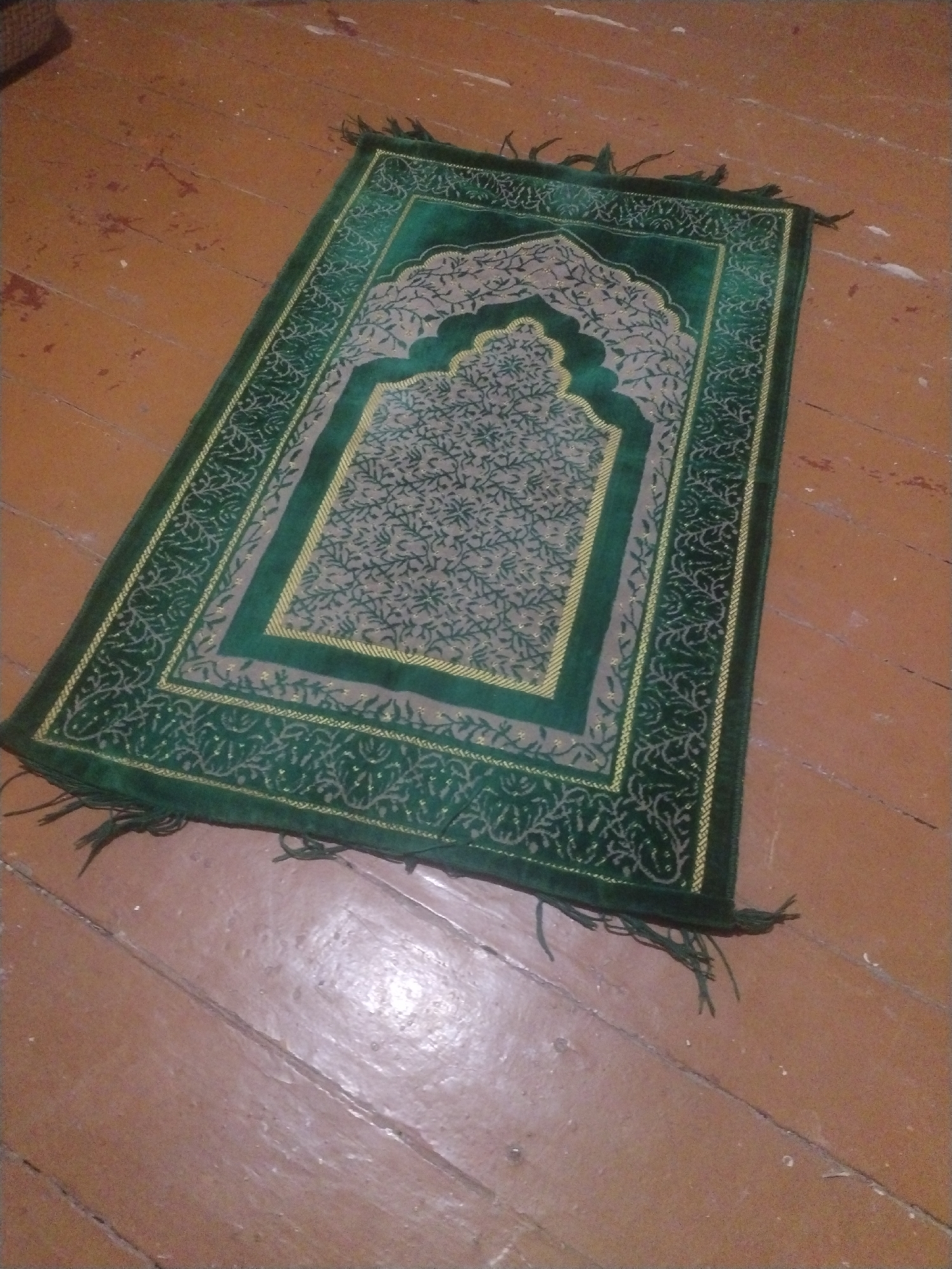 A green prayer rug with gold and beige accents is laid out on a wooden floor. The rug has a patterned design and fringe on the edges. The floor appears to be made of planks and has a worn, reddish-brown color. There is some light reflecting on the floor, indicating a light source from the left. The scene is viewed from a slightly elevated angle.
