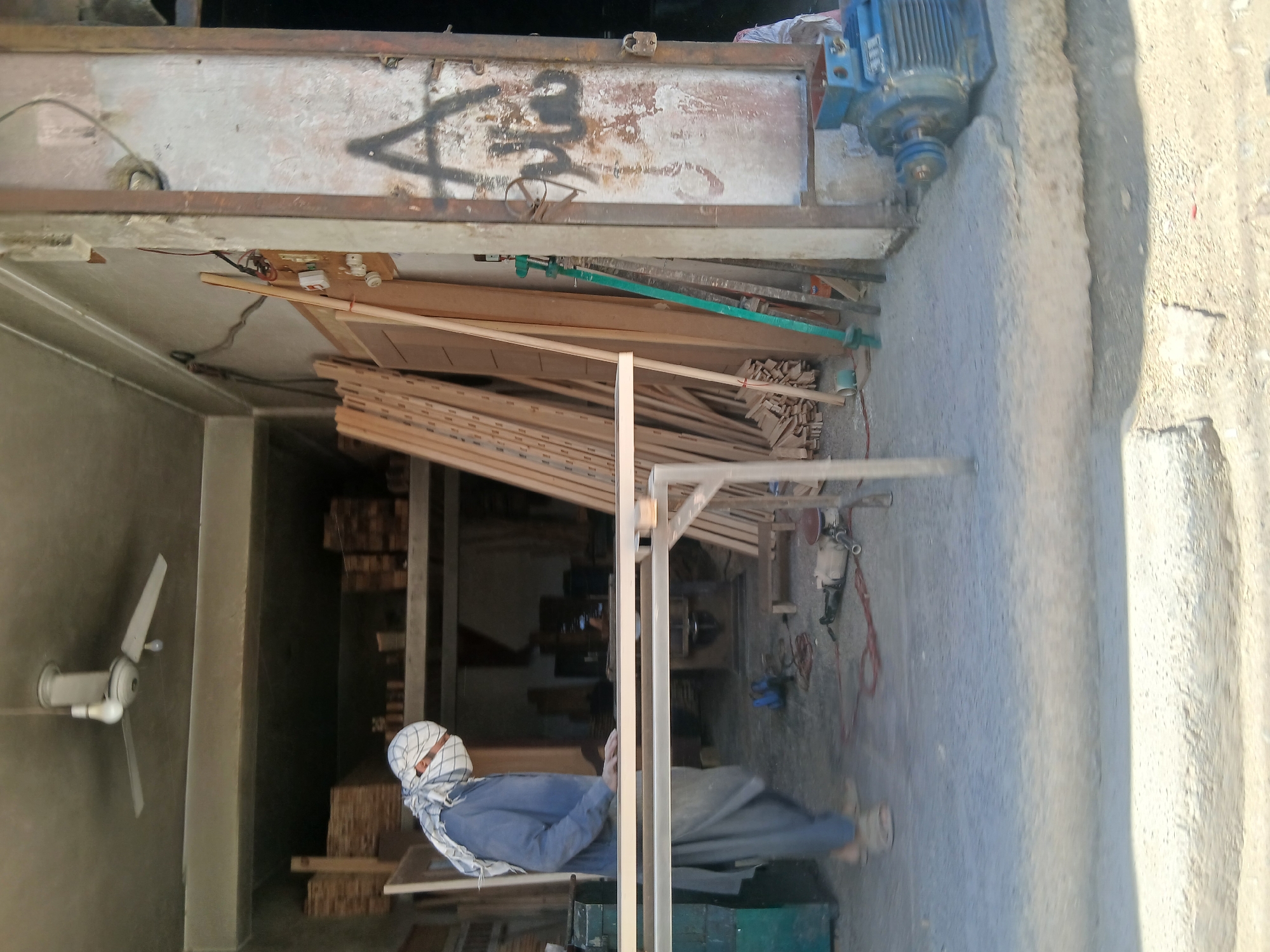 This media file captures an open-fronted workshop or carpentry shop in Quetta City Tehsil, Pakistan, viewed from just outside the entrance. The image is rotated 90 degrees counter-clockwise from its natural orientation; the description below refers to an upright view.
Inside the shop, the primary subjects are numerous stacks of wooden planks and smaller wooden pieces, indicating a woodworking trade. A person is visible within the shop, seated or bending, wearing a blue shirt and trousers, along with a white and blue checkered head covering that covers their face. To the left, a wall-mounted white fan is installed. Various tools are present, including a long green clamp or bar tool, and a power tool (possibly a sander or grinder) with red wiring is seen on the concrete floor. A light-colored wooden frame or stand is positioned in the foreground.
Above the entrance, a weathered metal shutter or door is prominent, displaying rust and graffiti. The graffiti includes a distinct, dark, angular symbol resembling an 'A' or an upward-pointing triangle, along with other indeterminate markings. Beneath this shutter, electrical components such as switches, wiring, and light sockets are affixed to a wooden board.
To the right of the entrance, outside the shop, there is a concrete ground surface that transitions into rough, textured steps or a pathway. A large, blue industrial motor with a pulley attachment is placed on the concrete edge near the entrance. The bright, clear lighting suggests it is daytime, with strong shadows present. The scene depicts an active, functional workshop environment.