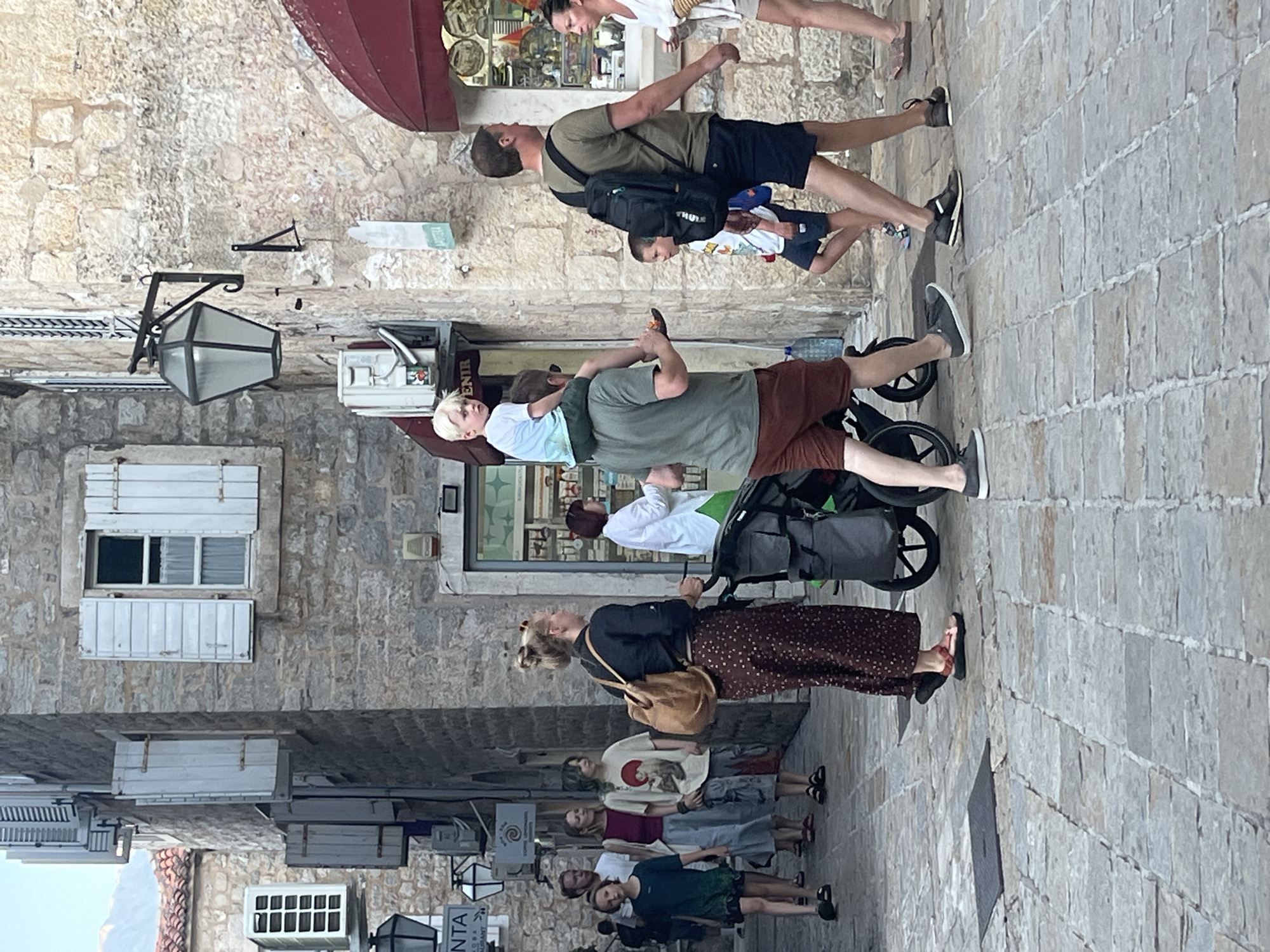 The image depicts a street scene in the historic old town of Budva, Montenegro, characterized by cobblestone pavement and stone buildings with traditional shutters. A group of people are walking along the narrow street.
In the foreground, a man wearing a green t-shirt and brown shorts walks on the right, with a young boy in a blue shirt and dark shorts walking beside him. Behind them, another man in a green t-shirt and dark shorts carries a child on his shoulders. This child is wearing a light blue t-shirt.
To the left of the main group, a woman with blonde hair pulled up in a bun, wearing a dark top and a patterned skirt, pushes a stroller. In front of her, a group of people are walking away from the camera. These include a person in a dark t-shirt and shorts, followed by another in a burgundy top and grey shorts, and then a person in a white t-shirt with a graphic print and camouflage shorts. Further back, more people are visible walking in the same direction.
The buildings flanking the street are constructed of light-colored stone. One building on the right features an awning and a shop window displaying merchandise. The building on the left has multiple windows with white shutters, some open and some closed, and an air conditioning unit mounted on the exterior. A large lantern is attached to the wall of the building on the right. The overall atmosphere suggests a warm day with moderate pedestrian traffic.