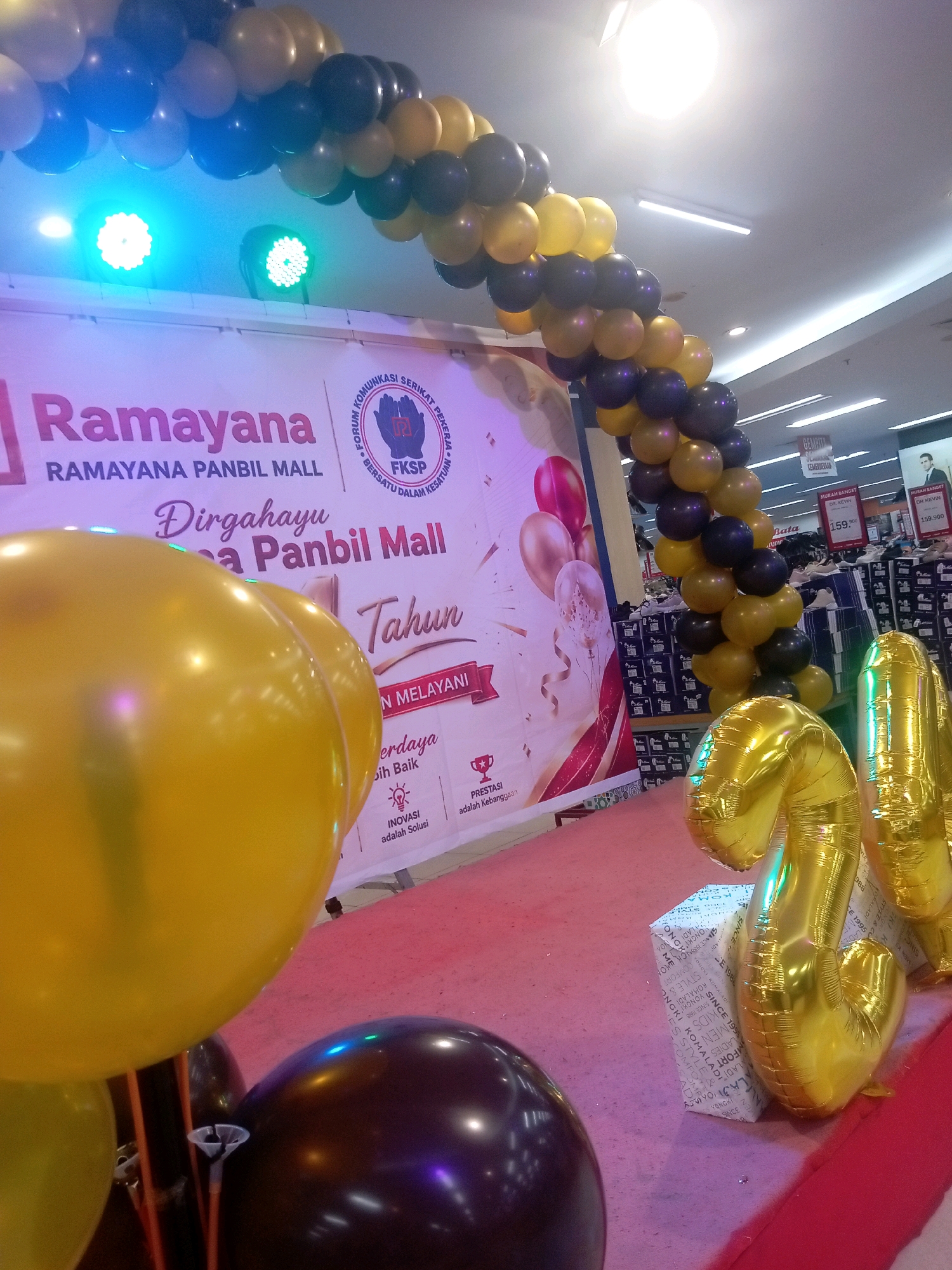 An indoor mall environment, likely Ramayana Panbil Mall in Batam, Indonesia, is decorated for a celebration. A large banner is visible with text in Indonesian, including "Ramayana Panbil Mall" and "Dirgahayu." An archway made of black and gold balloons spans across the top of the frame. In the foreground, large yellow and dark purple balloons are present. To the right, two large, shiny gold balloons shaped like the numbers "2" and "4" are standing upright on a white gift-wrapped box. The floor is covered with a red carpet. In the background, shelves of products and price tags are visible, suggesting the location is a retail store within the mall. There are no visible people in the frame.