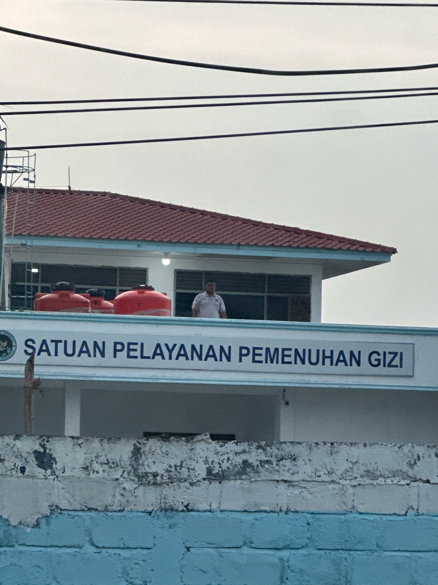 The image captures the facade of a building with red-tiled roofing. In front of the building, there are three large, red water tanks. A man in a light-colored shirt stands on the upper level of the building, near a window. A sign with blue text that reads "SATUAN PELAYANAN PEMENUHAN GIZI" is prominently displayed on the lower section of the building's facade. The building appears to be situated in an urban or semi-urban setting, as indicated by the presence of overhead power lines. The lower portion of the image shows a blue brick wall with weathered white plaster. The sky is overcast. This location is identified as being in the City of Binjai, Indonesia.
