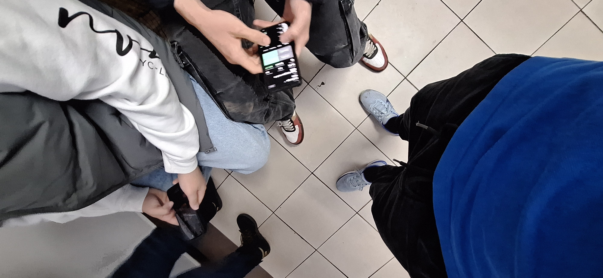 The image shows a group of four people standing in a public space, likely a subway or train station, based on the tiled floor. The image is taken from a high-angle perspective, looking down on the group.
The first person (from left to right) is wearing a white sweatshirt with black lettering, a grey jacket and blue jeans. The second person is wearing dark washed jeans, a dark jacket. They are holding a smartphone with a screen illuminated. The third person is wearing black sweatpants and pale blue running shoes. The fourth person is wearing a black jacket and bright blue sweatshirt.
All individuals are looking down, possibly at the smartphone being held by the second person.
The setting is an indoor location with square tiled floors. The walls are off-white. The scene appears to be well-lit.
There is no information in the image to suggest this image was taken in Burgas, Bulgaria.