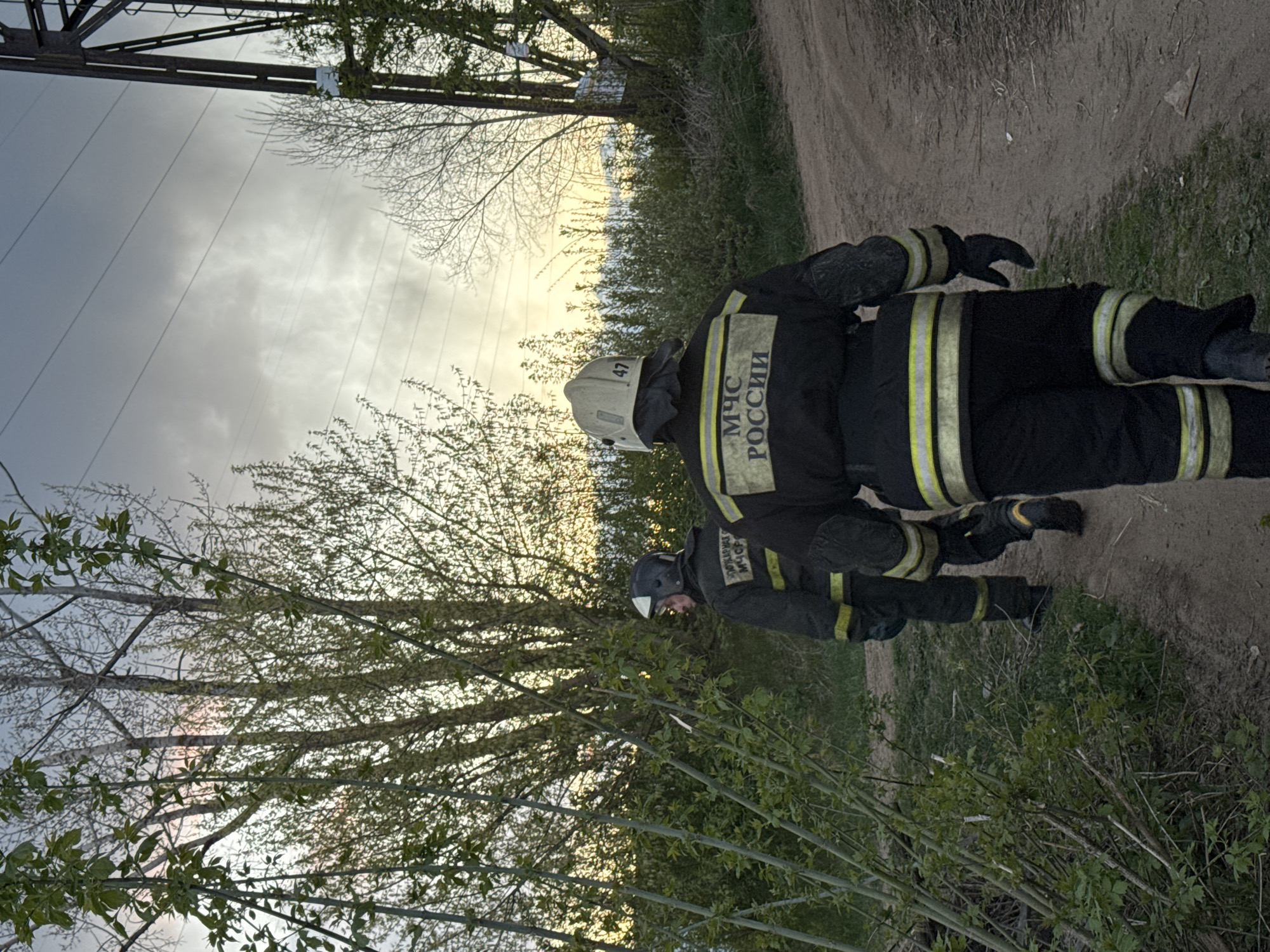 Two firefighters in black uniforms with yellow reflective stripes and helmets stand on a dirt path next to a grassy area with trees. The uniforms have "МЧС РОССИИ" (Ministry of Emergency Situations of Russia) printed on the back. The background shows a cloudy sky, power lines, and more trees.
While precise location within Chaikovsky cannot be confirmed with certainty from the image, the presence of "МЧС РОССИИ" and the general appearance of the scene indicate Russia.