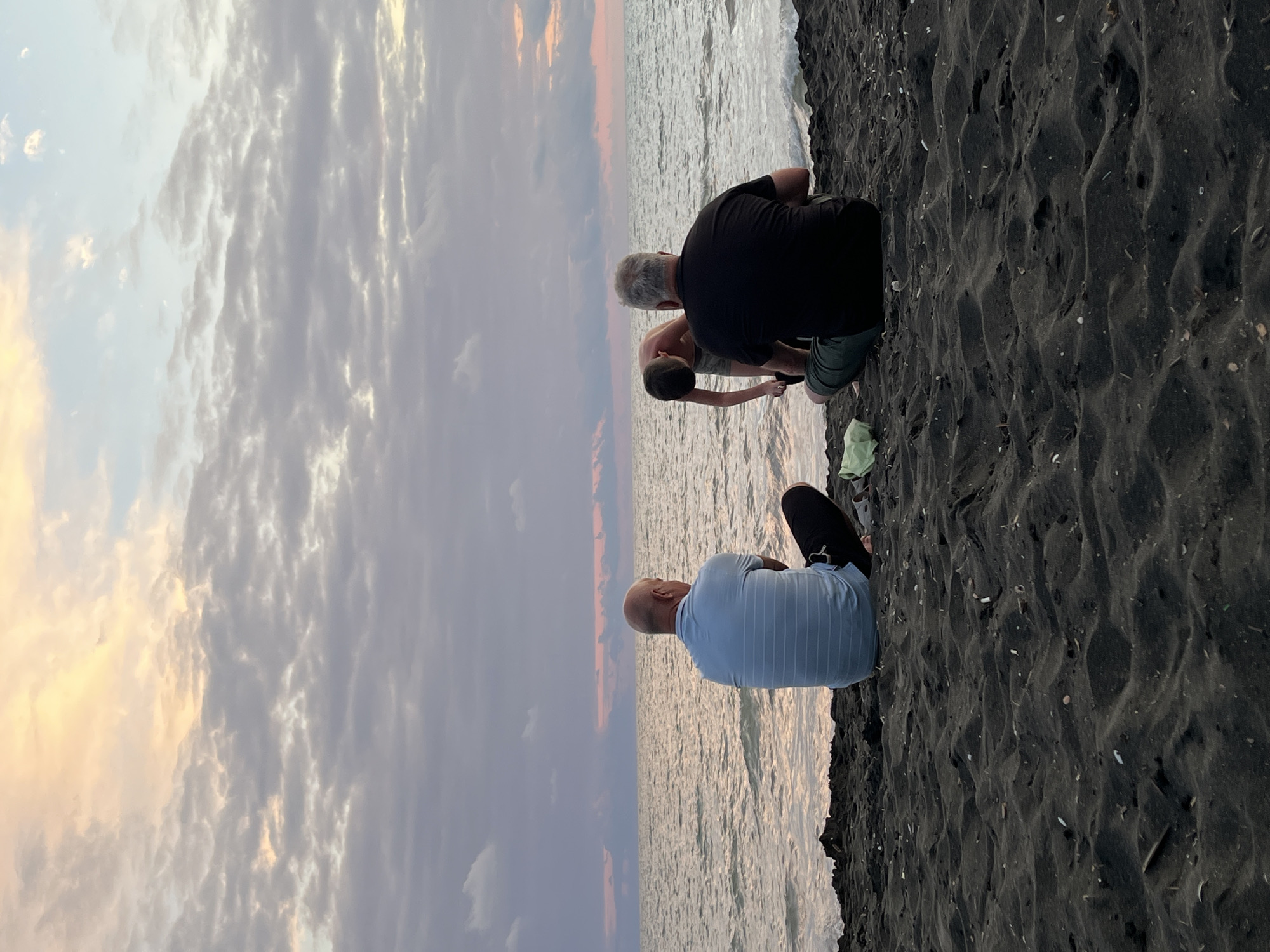 In Ureki, Georgia, three people are seated on a dark, sandy beach facing the Black Sea at dusk. The sky is filled with scattered clouds, tinged with hues of pink, orange, and purple. Two adult males are seated on the sand, facing the water. One, on the left, is wearing a light blue striped polo shirt and dark pants, and is sitting cross-legged. The other, on the right, is wearing a black t-shirt and green shorts, also sitting cross-legged. Between them, a young boy, shirtless and wearing gray shorts, is bent over, seemingly interacting with something on the sand or in the shallow water. A light green face mask lies on the sand to the left of the man in black. The sea is calm with small waves gently lapping at the shore. The dark sand is textured with ripples and dotted with small shells and debris.