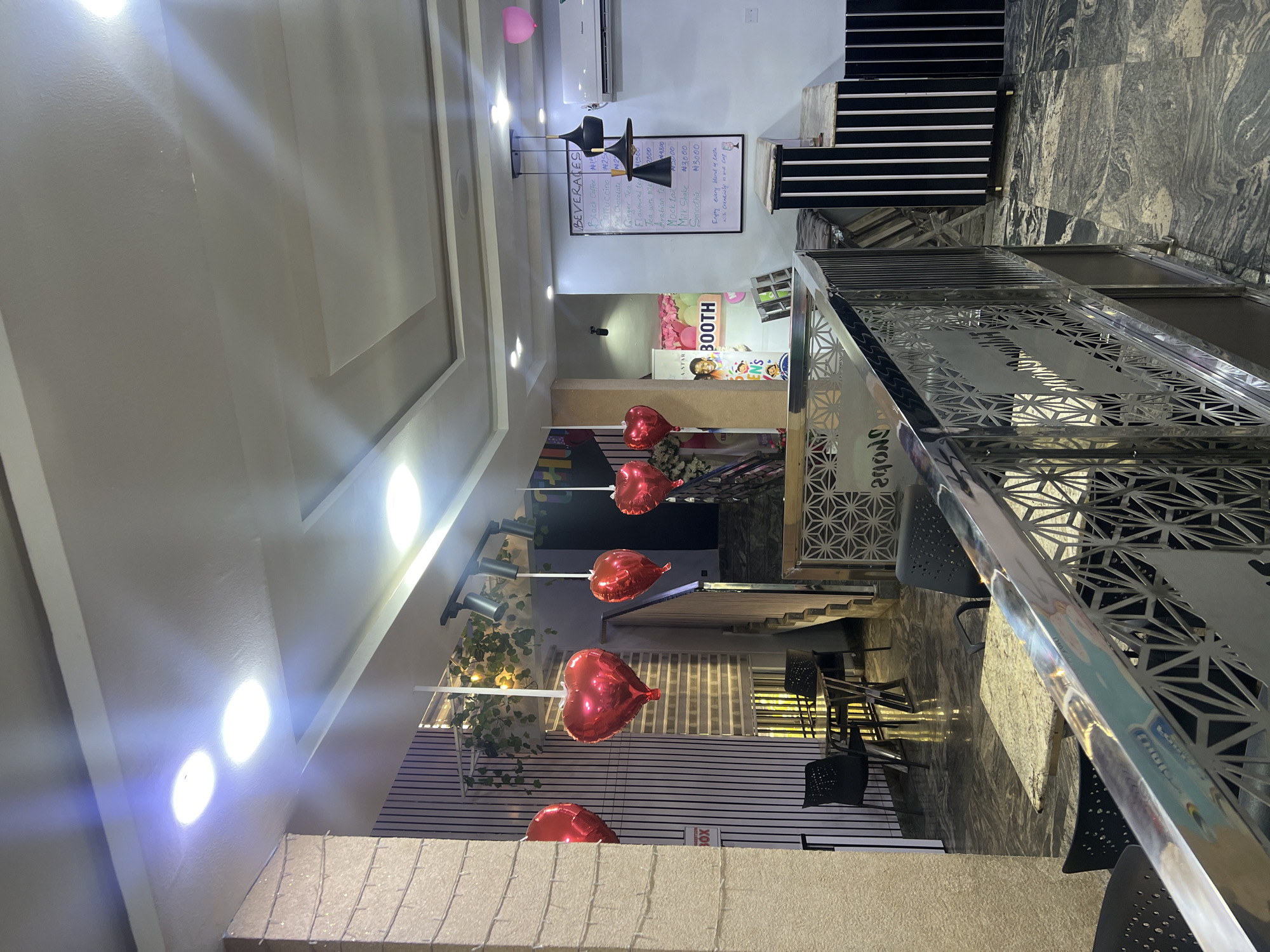 This is an indoor image of a cafe or restaurant. The scene is decorated with red heart-shaped balloons hanging from the ceiling, suggesting a celebratory or romantic atmosphere. In the background, there is a staircase and a booth area with a poster featuring a smiling child. A menu board displaying beverage options and prices is visible on the wall. The flooring appears to be polished stone with a marbled pattern. A metallic railing with an intricate design separates the foreground from the rest of the space. The lighting is bright, with overhead spotlights and pendant lamps. The overall impression is that of a modern and welcoming establishment.