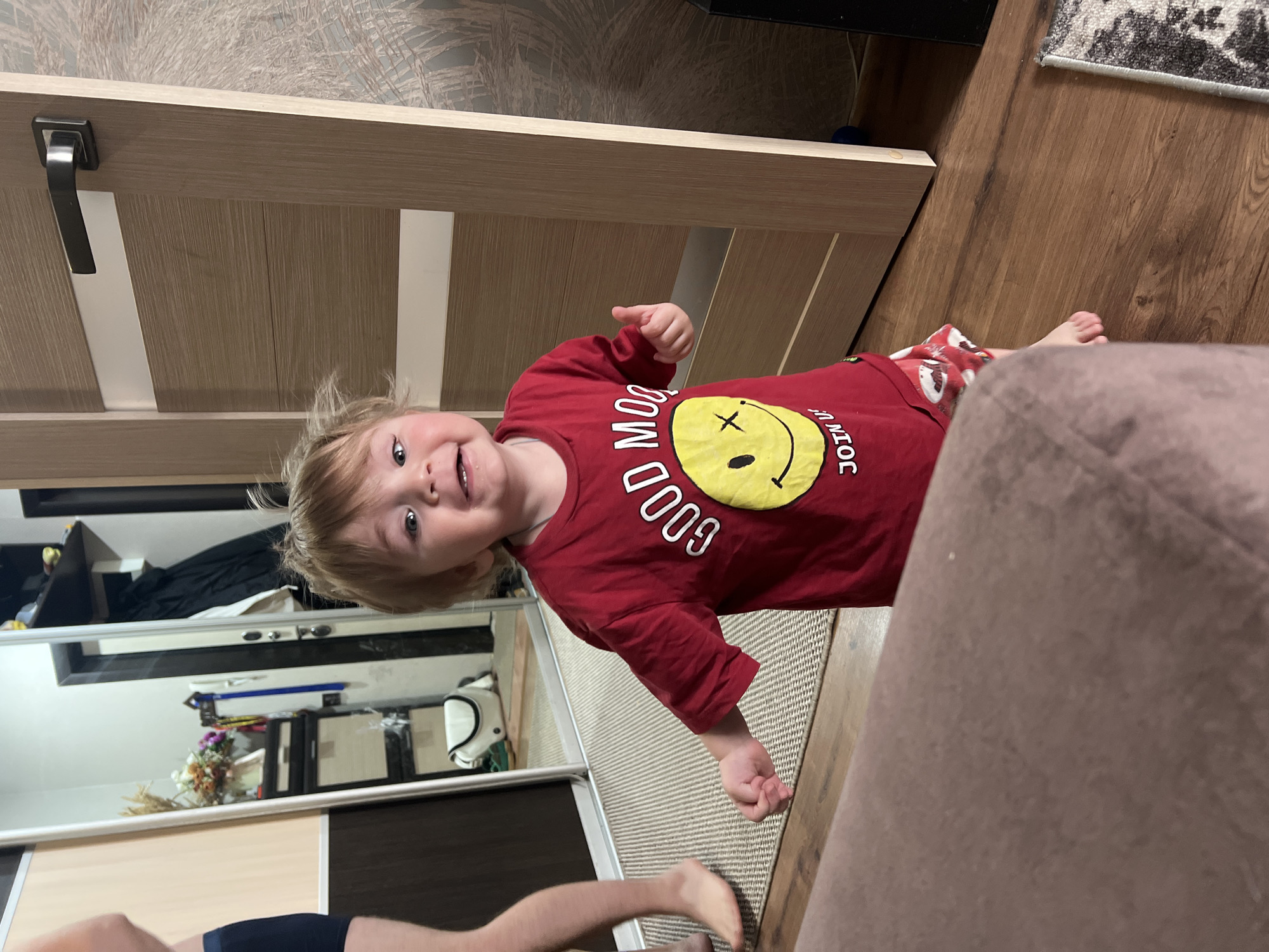 A toddler wearing a red long-sleeve shirt with a yellow smiley face graphic and red and white patterned shorts is looking toward the camera and pointing with their right hand. The setting appears to be an indoor living space. A wooden door is visible on the left, and a section of hardwood floor on the right. A person's leg and foot are visible at the bottom left, suggesting another person is present. The scene is likely taking place in a home, as indicated by the door, hardwood floor, and other indoor elements. Based on the information provided, the location is likely in the city of Noviy, Russia.