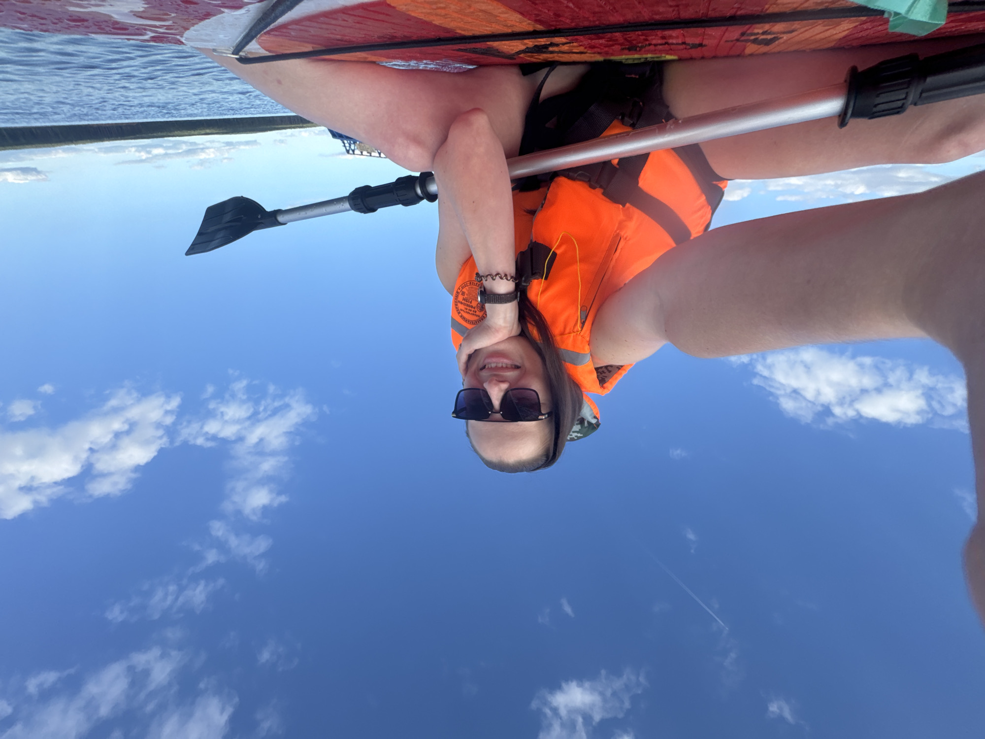 An outdoor, low-angle, medium shot shows a woman sitting on a paddleboard on a body of water. The woman, with her legs spread wide and touching the board, is wearing an orange life vest, black shorts, and sunglasses. She has one hand on her chin, with her other arm extended towards the camera. A paddle, with its blade slightly visible, is placed across her lap and the board. The water is calm, and a forest lines the horizon under a blue sky with scattered white clouds. The image appears to be taken on the Kama River in the city of Chaikovsky, Russia.