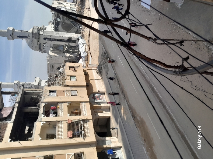 This image depicts a street scene in Khan Yunis, Palestinian Territory, viewed from an elevated perspective. The foreground features a rusty metal railing and numerous tangled electrical or communication wires. A paved road, partially marked with white lines and showing signs of wear, stretches through the midground. Multiple pedestrians, including adults and children, are walking on the road and along its edges.
To the left, a multi-story building, light brown in color, exhibits severe structural damage; its upper floors are partially collapsed with exposed interiors and rebar, while lower floors retain some balconies, one of which has clothes hanging. To the right and center-background, a large mosque with a central dome and two minarets stands significantly damaged. Both minarets show fractured sections, and the mosque's walls and dome have large sections missing or compromised, revealing internal structures. Rubble and debris are scattered across the ground between the road and the damaged mosque, with two white, tarp-covered structures visible amidst the wreckage. The sky is clear, suggesting daytime conditions. The overall scene is one of an urban environment heavily impacted by destruction, yet populated with daily activity.