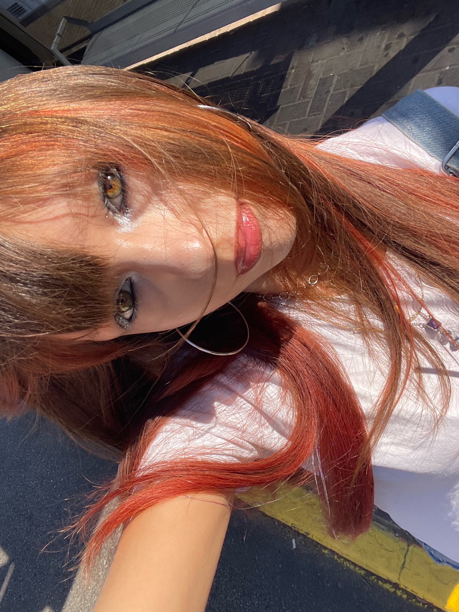 The image is a close-up, low-angle, outdoor shot of a young person with auburn hair featuring lighter, reddish-blonde streaks. The person is wearing a white t-shirt and a denim garment, possibly shorts or a skirt, with visible stitching and hardware. They are also wearing hoop earrings and necklaces. The person's face is partially obscured by their hair, but their eyes are visible, with makeup applied to the eyelids. Their lips are glossy and appear to be painted a pinkish-red color. The background consists of a paved surface with a yellow line, suggesting an urban environment, possibly a street or sidewalk. The lighting indicates it is daytime, with strong sunlight casting shadows. The image is oriented such that the person's head is towards the top left. There are no discernible landmarks or specific features that would pinpoint a location within the city of Raanana, Israel.