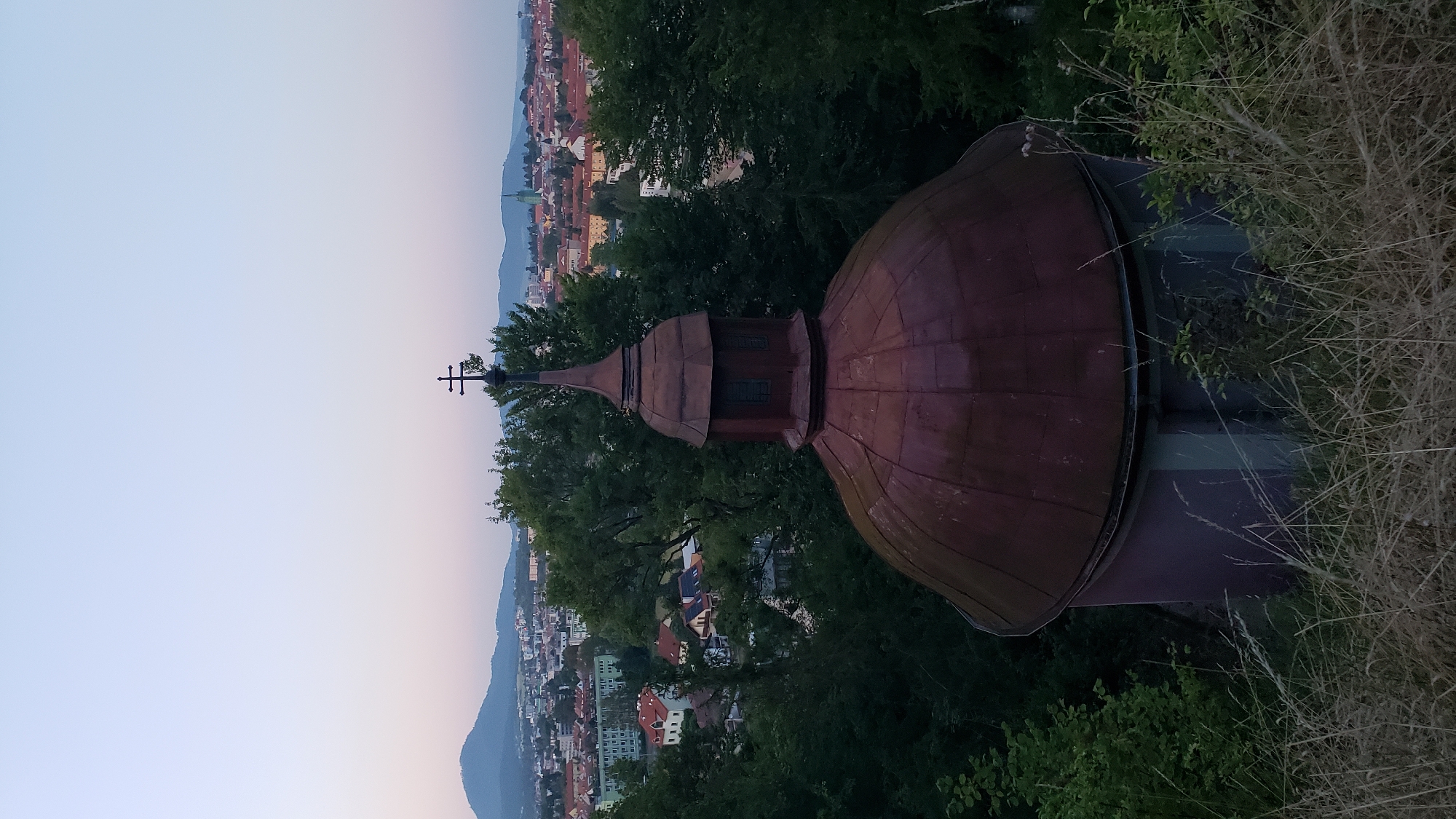 An exterior, medium shot captures a copper-domed structure, possibly a chapel or a turret, with a cross atop its spire. The dome is weathered and appears to be a deep reddish-brown. The structure is situated on a grassy hill with dry, tall grass in the foreground. Behind the dome, a panoramic view of the city of Prešov, Slovakia, unfolds. Numerous buildings, including residential houses with orange tile roofs and taller structures, are visible, interspersed with dense green trees. In the distance, rolling hills and mountains form the backdrop under a pale, twilight sky. The lighting suggests it is either early morning or late evening. No people are present in the frame. The scene appears to be a serene, elevated viewpoint overlooking the city.