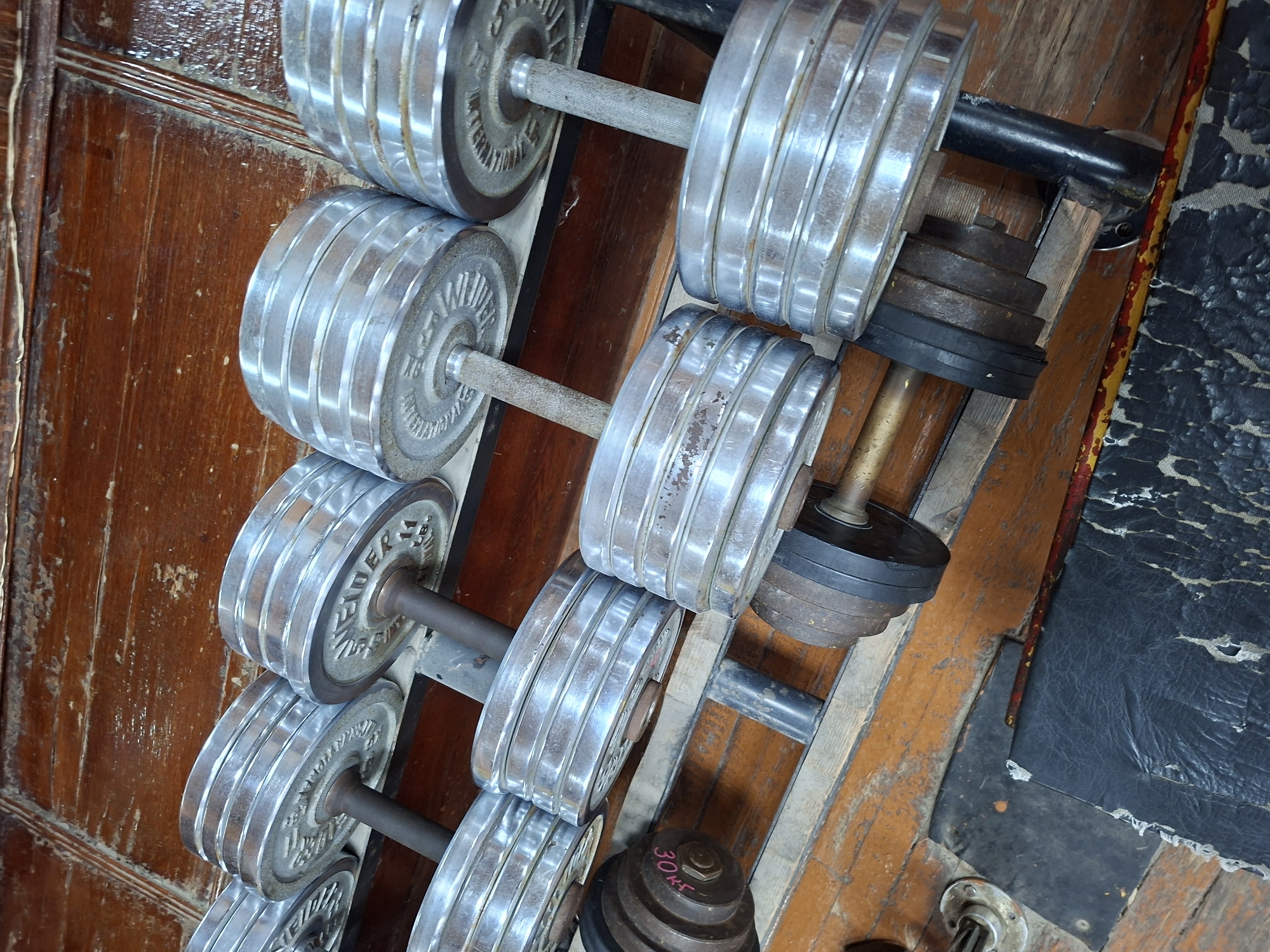 The image depicts a close-up, low-angle shot of a rack of weightlifting dumbbells. The dumbbells are made of metal and are stacked with circular weight plates. The rack itself appears to be made of wood, with a worn and aged finish. In the foreground, a black, cracked mat lies on a wooden floor. The scene is dimly lit, with the focus on the metallic sheen of the weights. The overall impression is one of a used and perhaps slightly neglected gym environment. There is no specific visual information to suggest a location in Kazan, Russia.