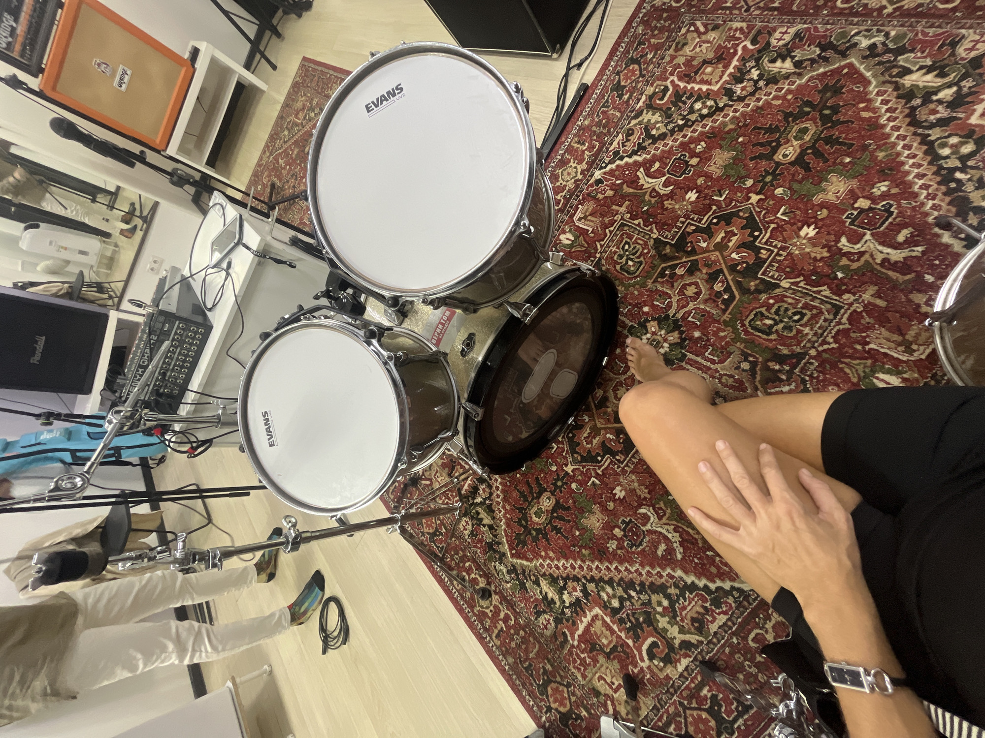 The image shows a drum set, a person's legs, and a recording studio environment. The drum set includes at least two drums with white drumheads, one of which has "EVANS" branding. A person's legs, wearing black shorts and a watch, are visible in the foreground. They are sitting on a red and black patterned rug. In the background, there's a mixing board, a microphone stand, and a large orange amplifier. The room appears to be a rehearsal or recording space. There is no visible indication of the city being Moscow, Russia.