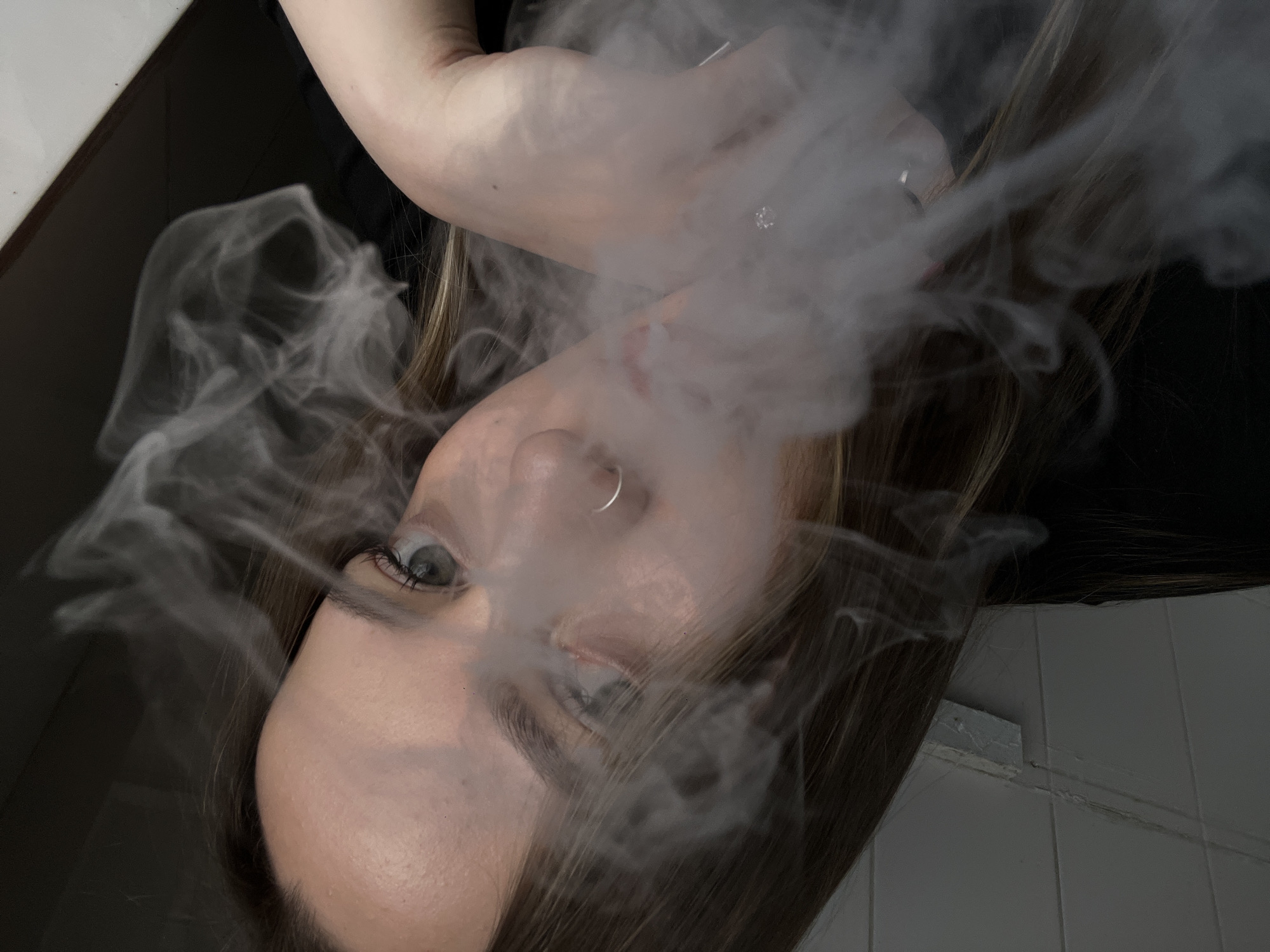 A woman with fair skin, light brown hair, and a silver nose ring is seen exhaling a cloud of white vapor. Her head is oriented with her right cheek facing downwards, and her gaze is directed upwards and to the left. She is wearing a black top. Part of her arm and hand with a ring on one finger are visible above her face in the top portion of the frame. The background includes a white tiled wall and what appears to be a dark countertop or table. The location is not discernable from the visual information in the image.