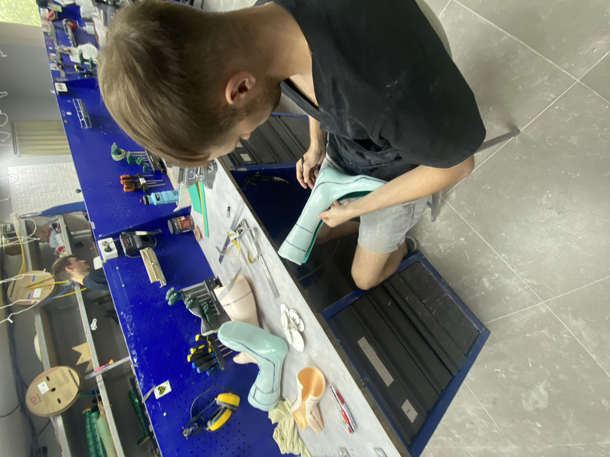 In a workshop, a young man with brown hair and a black t-shirt is sitting and working on a light blue prosthetic leg. He is holding a tool and appears to be marking the prosthetic. On the table in front of him are other prosthetic leg parts, tools, a measuring tape, and a red pen. In the background, a blue pegboard is filled with various tools. Another person is visible in the distance, also working in the workshop. The environment appears to be a professional setting for creating prosthetics.