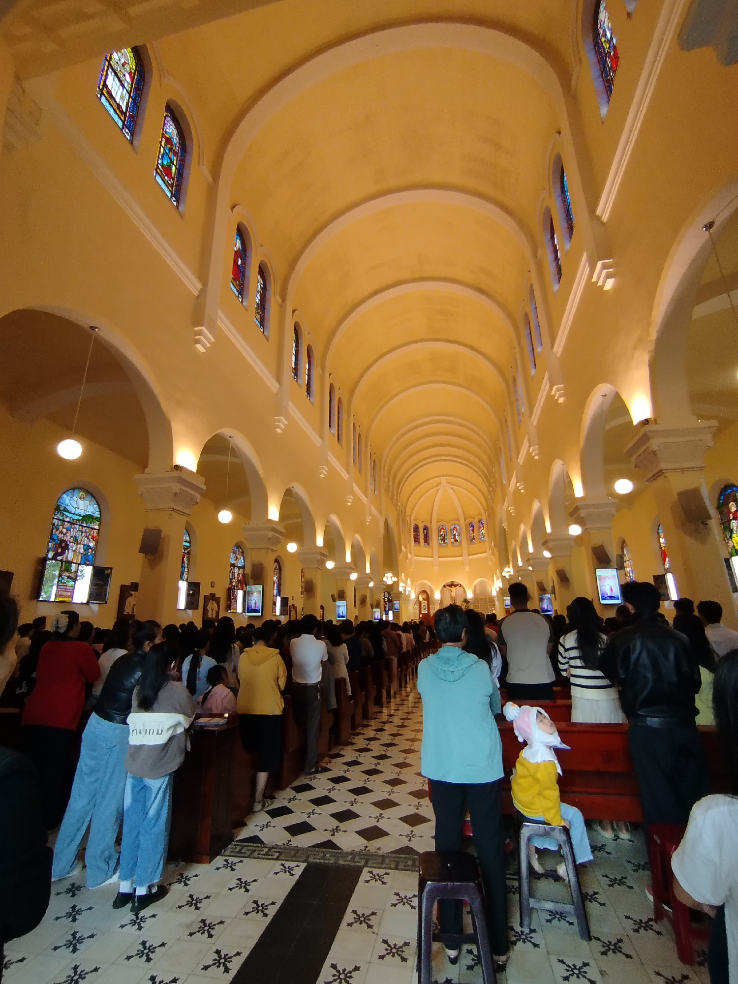 The image depicts the interior of a church in Da Lat, Vietnam, filled with people. Rows of individuals are standing or kneeling in the pews, facing towards the altar at the far end of the building. Arched ceilings and stained-glass windows characterize the architecture. The floor is covered in a black and white patterned tile. A child wearing a rabbit hat is seated on a stool in the foreground.