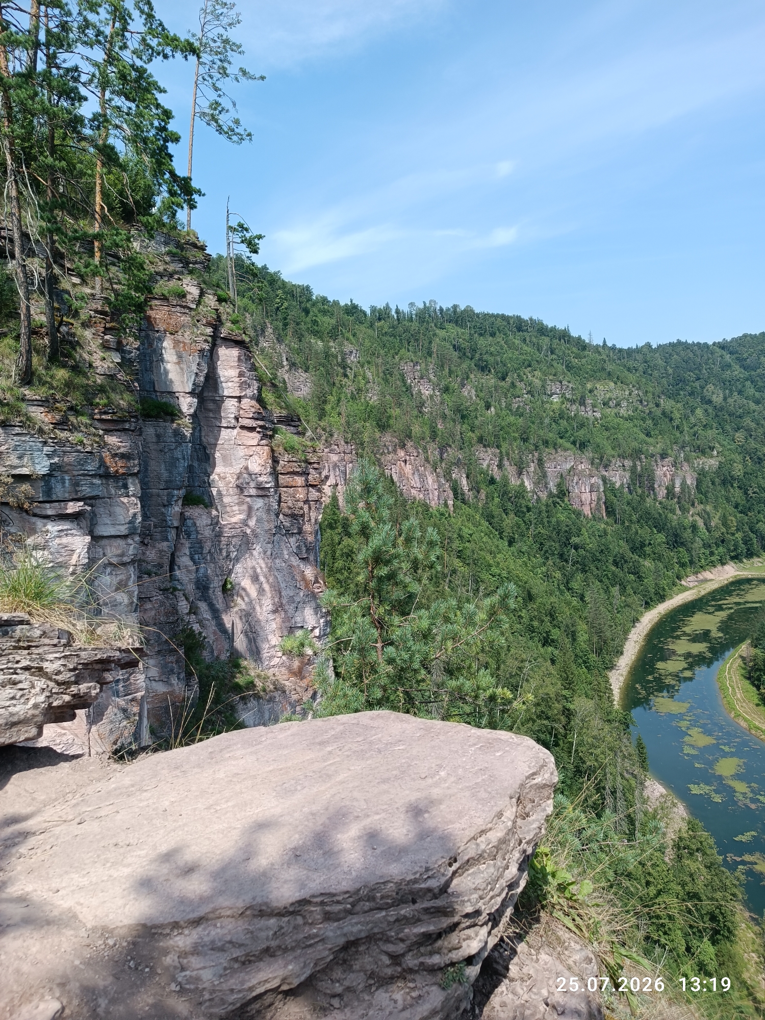The image shows a scenic view of a rocky cliff face with a winding river below. The cliff is covered in dense green trees and vegetation. The river is a dark blue-green color and has some patches of green algae floating on its surface. A dirt path runs along the edge of the river. In the foreground, a large, light-colored rock formation dominates the lower portion of the image. The sky is blue with a few scattered white clouds. The date stamp in the lower right corner reads "25.07.2026 13:19". The location is identified as Brishtamak, Russia. There are no people or animals visible in the image. The overall impression is one of a natural, untouched landscape.