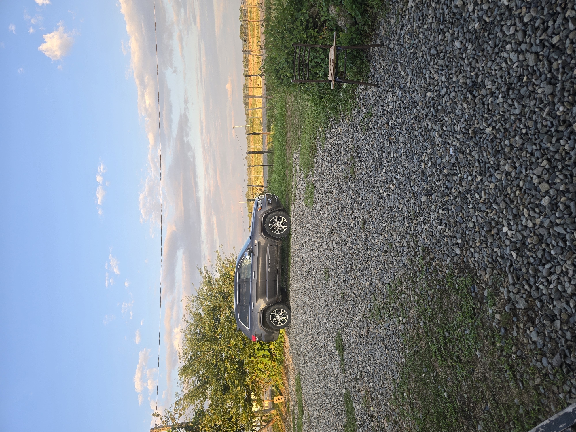 A dark gray Jeep Grand Cherokee is parked on a gravel driveway next to a dense green tree. The driveway is covered in small, dark gray and tan pebbles. To the left of the car, the tree's foliage is thick and lush, with sunlight filtering through its branches. In the distance, a fence line runs horizontally across the frame, with a field of vegetation beyond it. The sky is a bright blue with scattered white and wispy clouds, tinged with warm hues of orange and pink, suggesting it's either early morning or late afternoon. A thin black wire stretches across the sky, likely a utility line. A metal chair with wooden slats is placed near some dense green bushes in the background. The overall setting appears to be a rural or semi-rural area in Ingiri, Georgia. There is no visible text in the image.