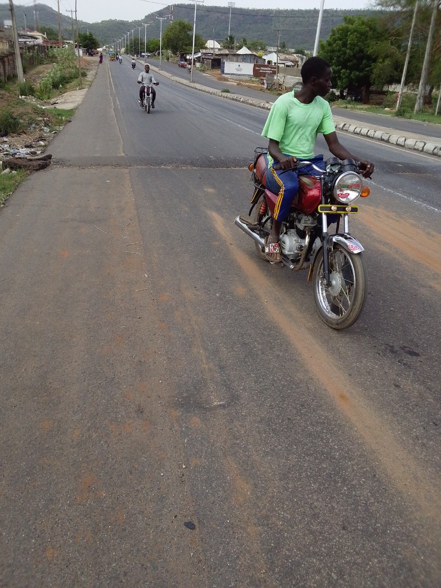 The image shows a wide, paved road in Adankolo, Nigeria. Two individuals are riding motorbikes. The closest motorbike is ridden by a Black male wearing a light green T-shirt and blue pants with yellow stripes. He is looking to his left. The other motorbike is further down the road and the rider is wearing lighter clothing. The road has some debris on the shoulder and an uneven section where it meets the roadside. There are buildings and trees lining the road. Mountains are visible in the distance. Overhead power lines and streetlights are present.
